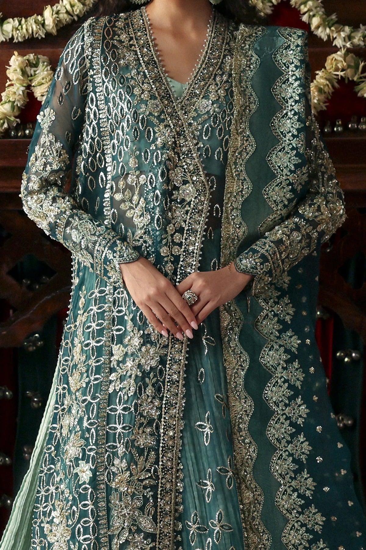A close-up of a woman wearing an ornate teal green embroidered traditional outfit with intricate silver and gold metallic detailing, paired with a matching sheer dupatta.