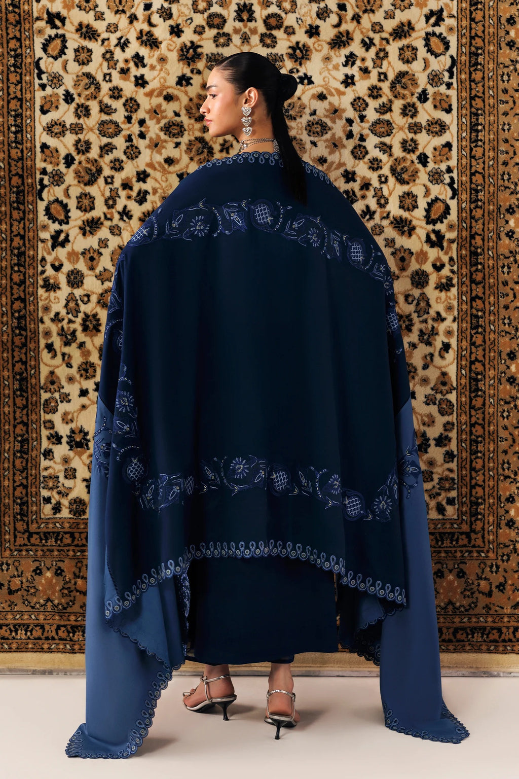 A woman stands with her back to the camera wearing a large navy blue shawl with intricate light blue floral embroidery, set against a patterned beige and brown rug.