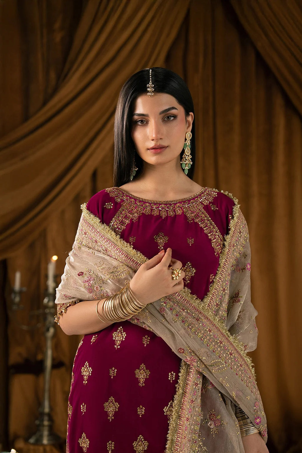 A woman models a traditional Indian outfit featuring a deep magenta embroidered kurta with a beige dupatta draped over her shoulders, set against a warm brown draped fabric background.