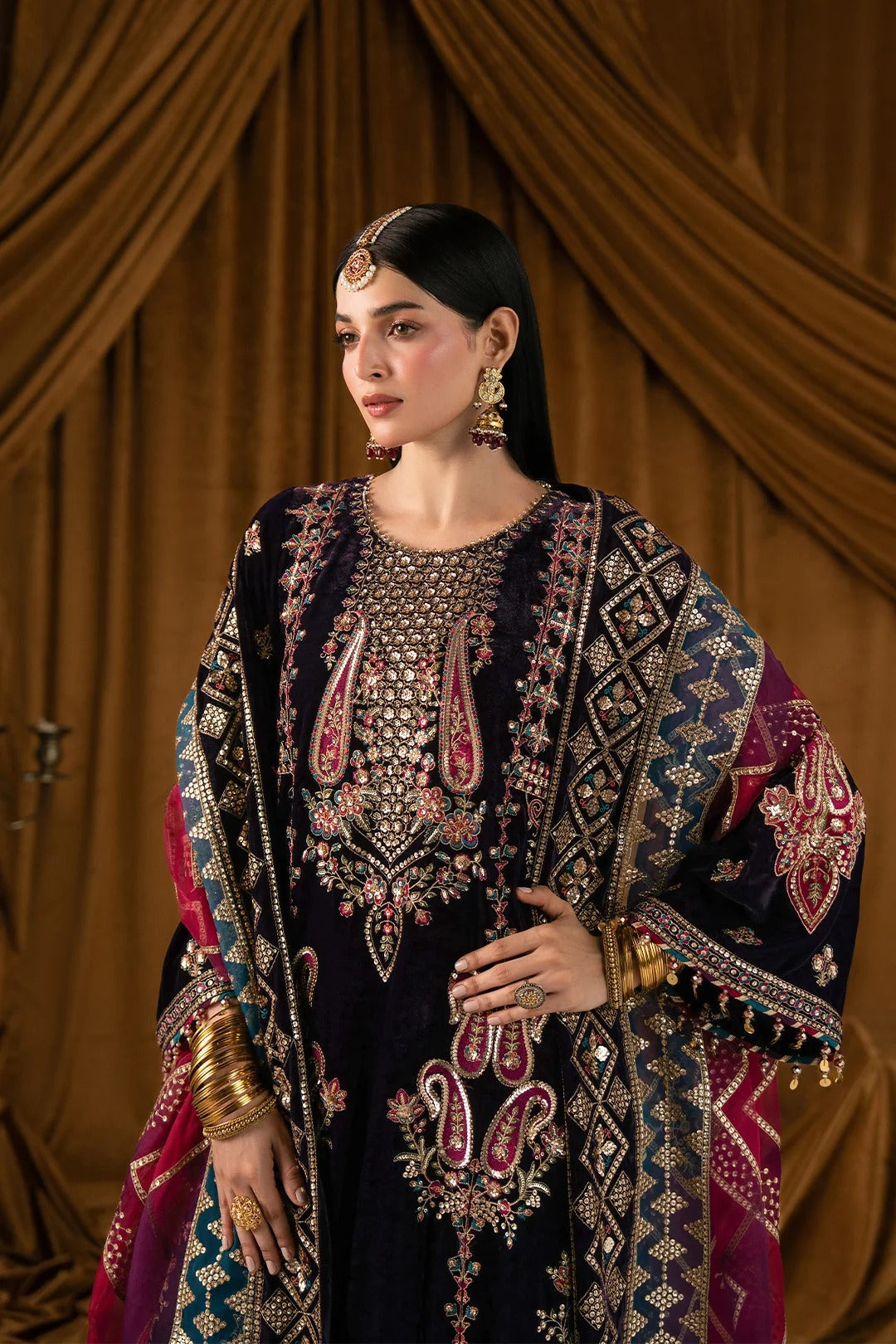A woman models an ornate dark navy traditional outfit with extensive gold embroidery, pink paisley accents, and a matching patterned dupatta, set against a draped brown fabric backdrop.