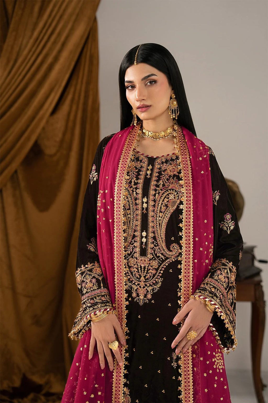 A woman modeling a traditional black and magenta ethnic outfit with intricate gold embroidery, a matching magenta dupatta, and gold jewelry, set against a studio backdrop with a brown draped curtain.