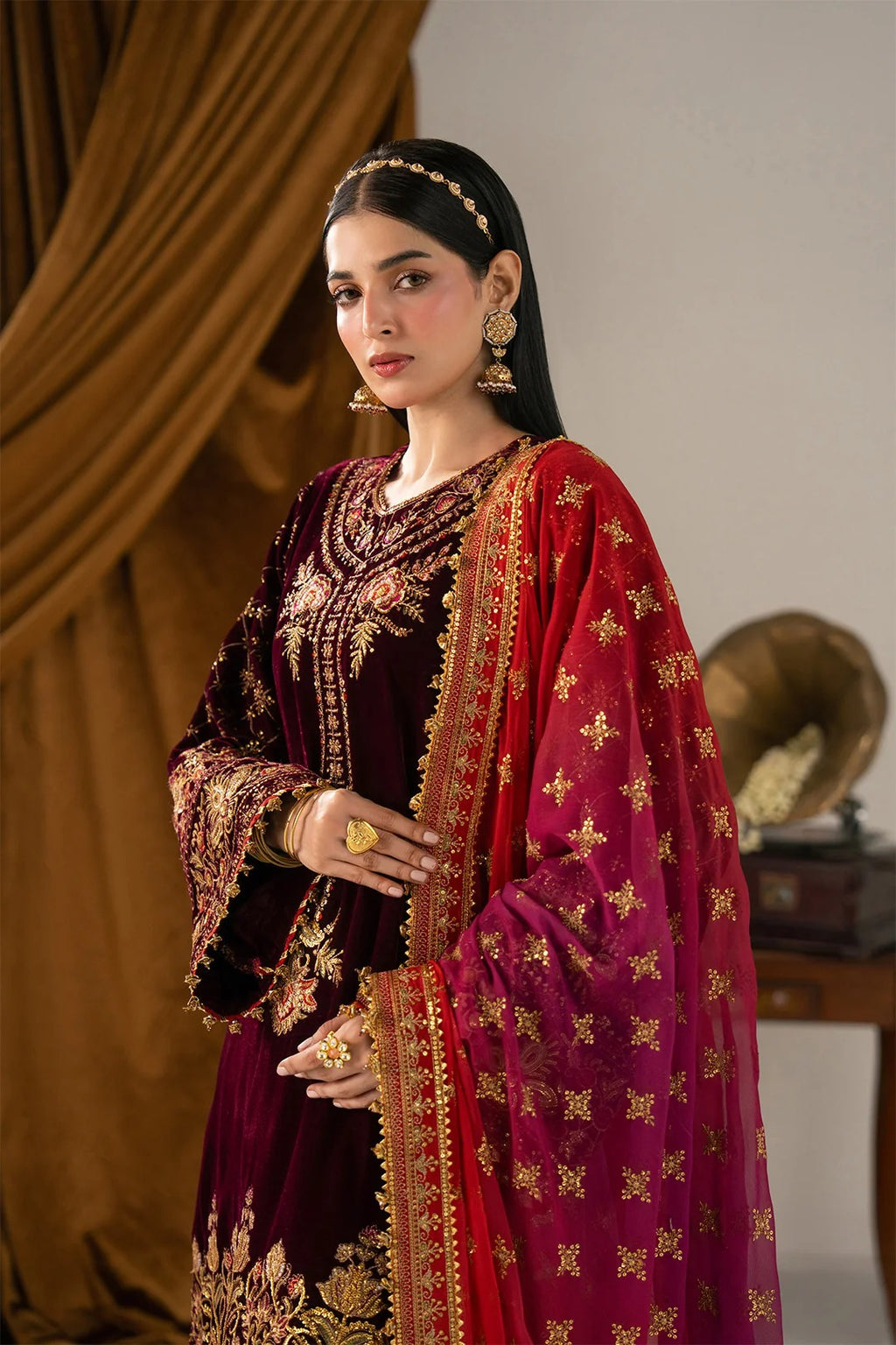 A woman models an ornate traditional Indian outfit featuring a deep maroon velvet kurta with intricate gold embroidery and a contrasting magenta dupatta decorated with gold motifs, set against a warm brown draped background.