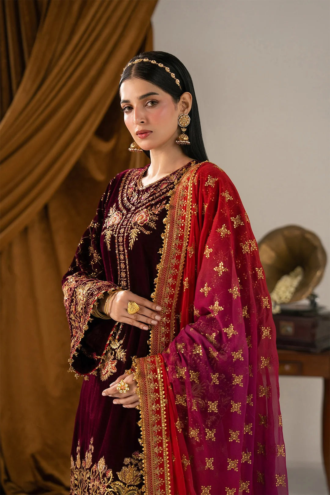 A woman models an ornate traditional Indian outfit featuring a deep maroon velvet kurta with intricate gold embroidery and a contrasting magenta dupatta decorated with gold motifs, set against a warm brown draped background.
