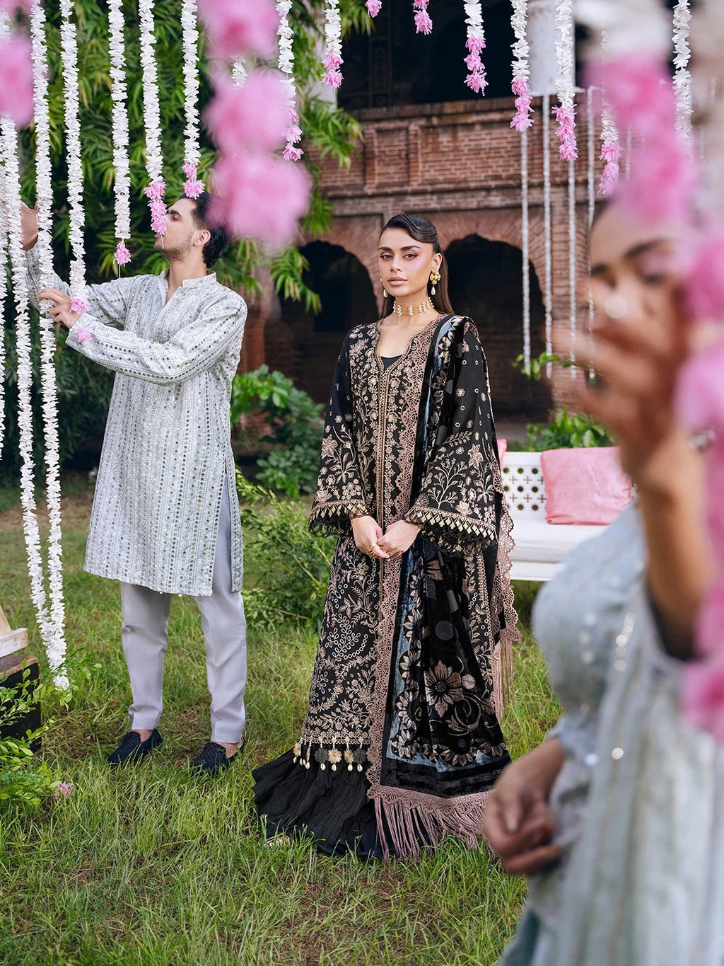 A woman in an ornate black and gold embroidered traditional outfit stands outdoors on grass, surrounded by pink hanging flower garlands and a man in a light gray kurta adjusting the garlands. Another person is partially visible in the foreground, and a brick building is in the background.