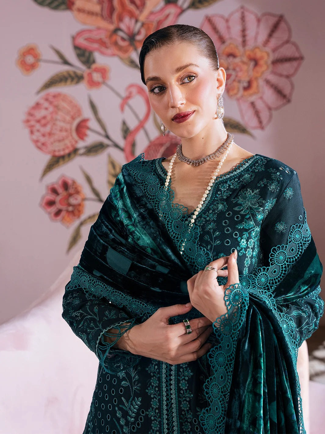 A woman wearing a dark teal green velvet embroidered shawl and matching embellished top, posed against a pink floral wall backdrop.