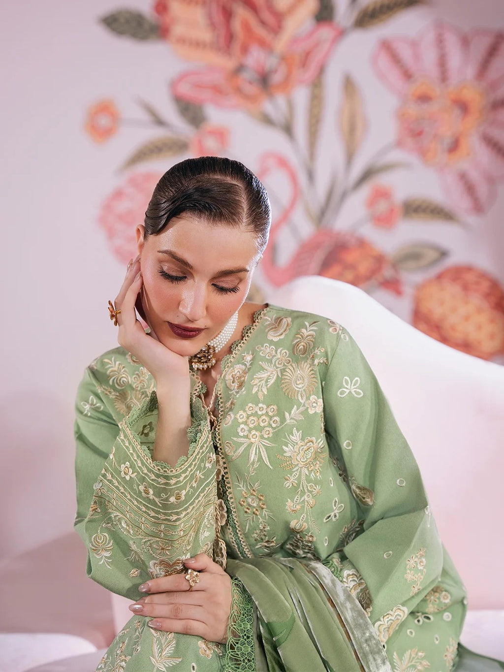 A woman wearing a sage green embroidered traditional outfit with gold detailing, seated against a pink floral wall and white chair.