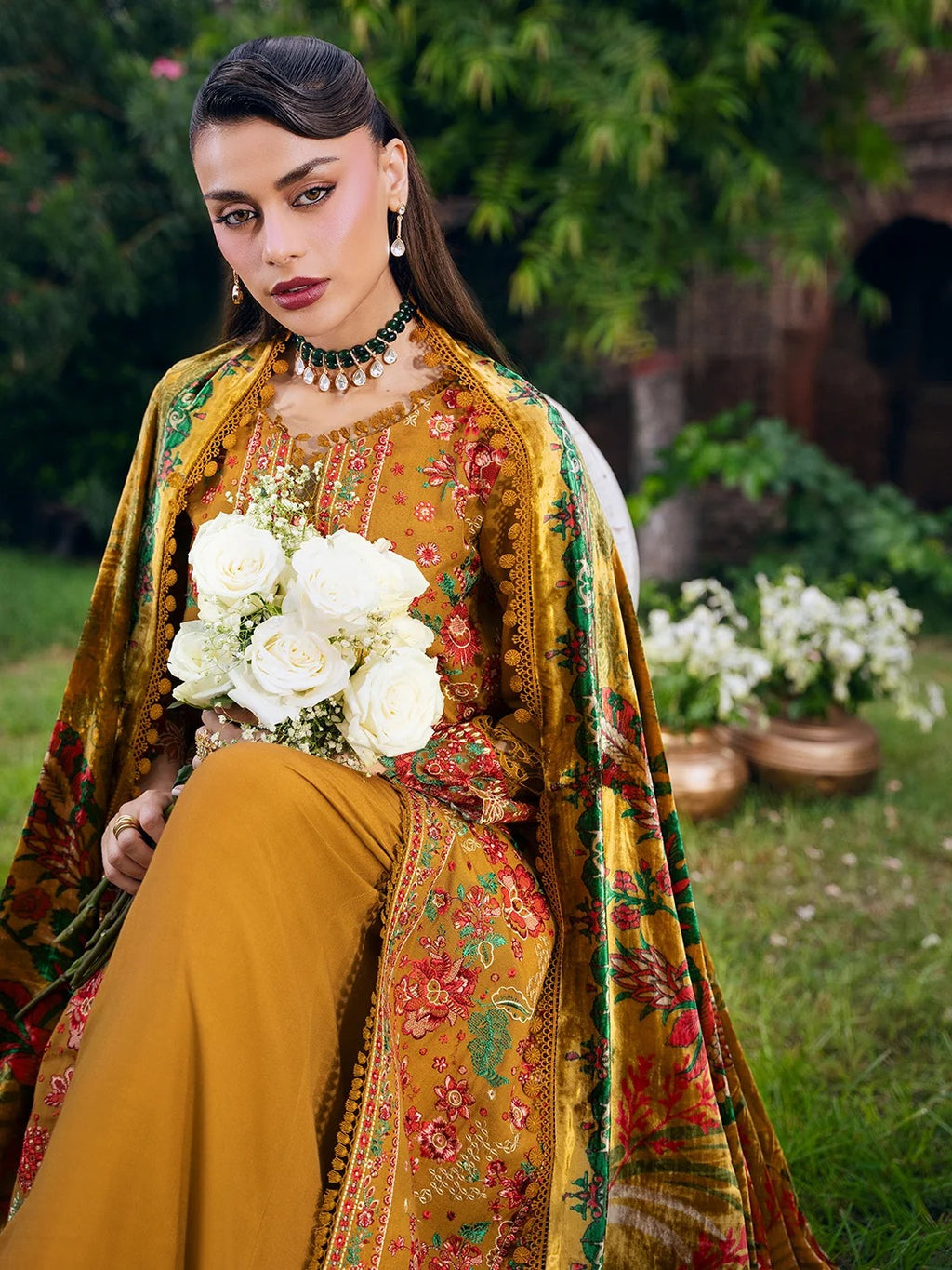 A woman stands outdoors in a garden setting wearing a traditional embroidered outfit. The outfit is a mustard yellow three-piece ensemble with intricate floral embroidery in red, pink, and green, and a matching dupatta draped over one shoulder.