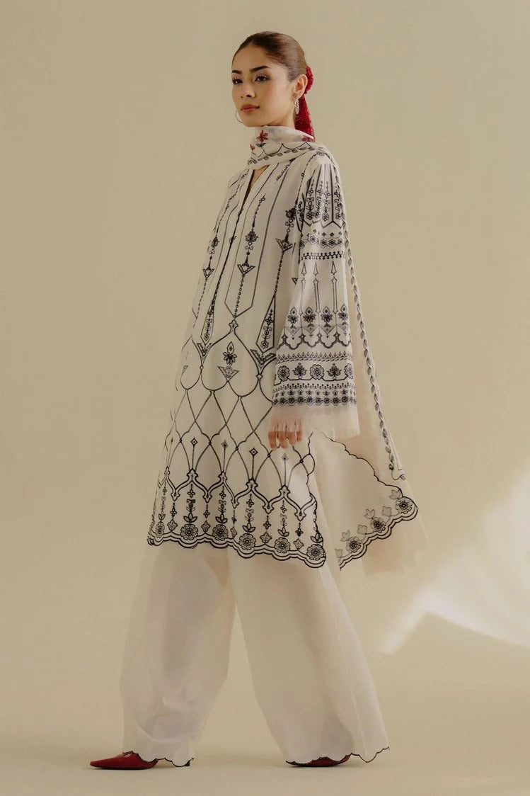 A model wearing a white traditional outfit with black geometric embroidery, featuring a high-neck tunic, wide-leg pants, and a matching dupatta, set against a solid beige background.