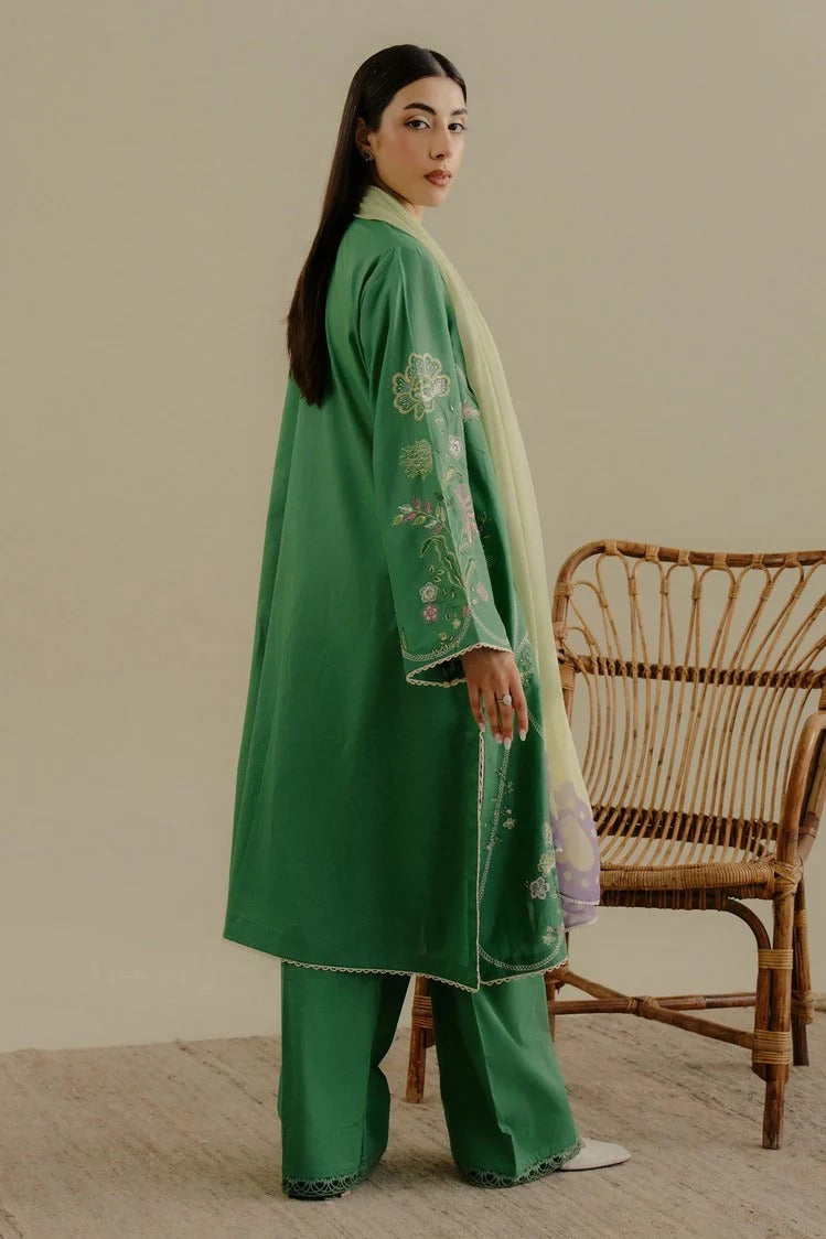 A woman models a green traditional outfit consisting of a long kurta with floral embroidery on the sleeve, matching wide-leg trousers, and a light green dupatta, standing next to a wicker chair against a beige wall.