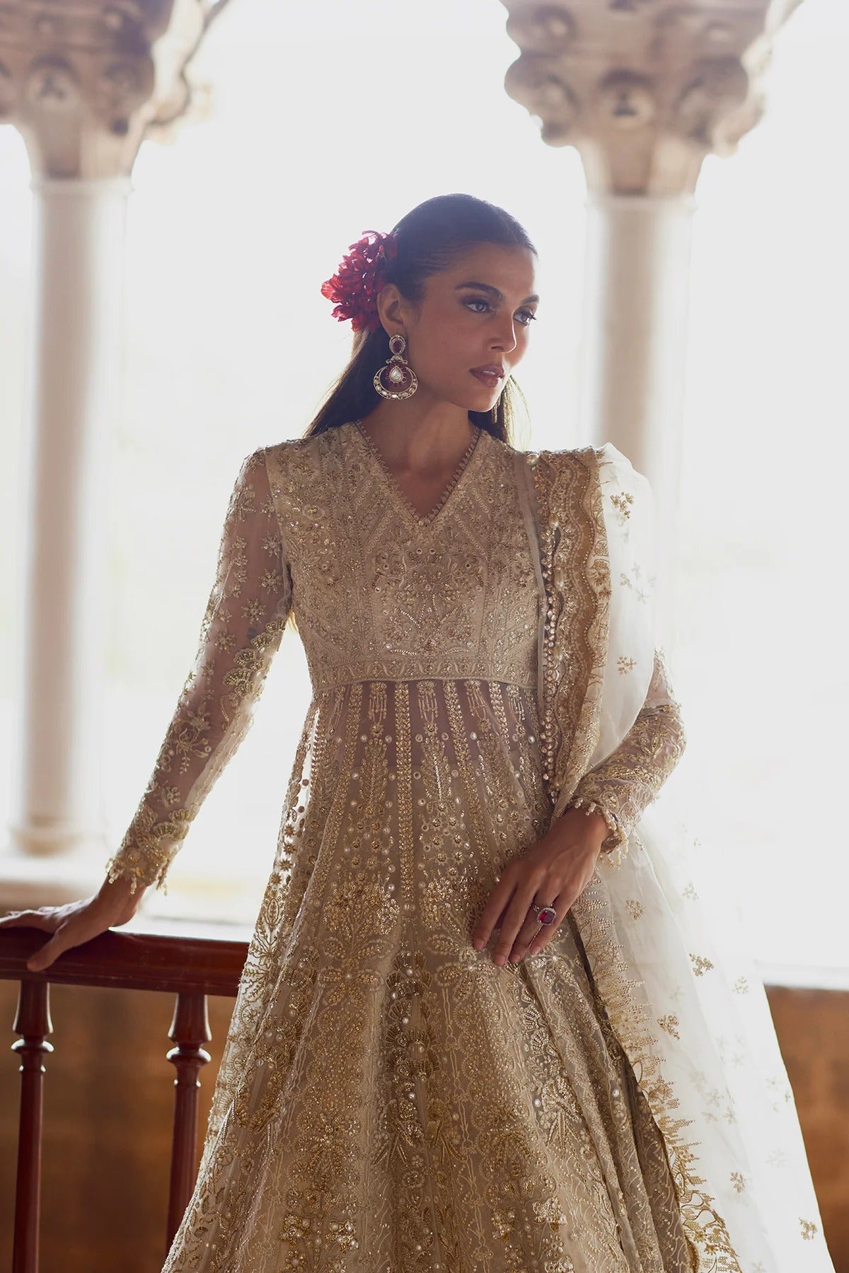 A woman wearing an ornate beige and gold embroidered traditional dress with a white dupatta, standing indoors near columns and a railing.