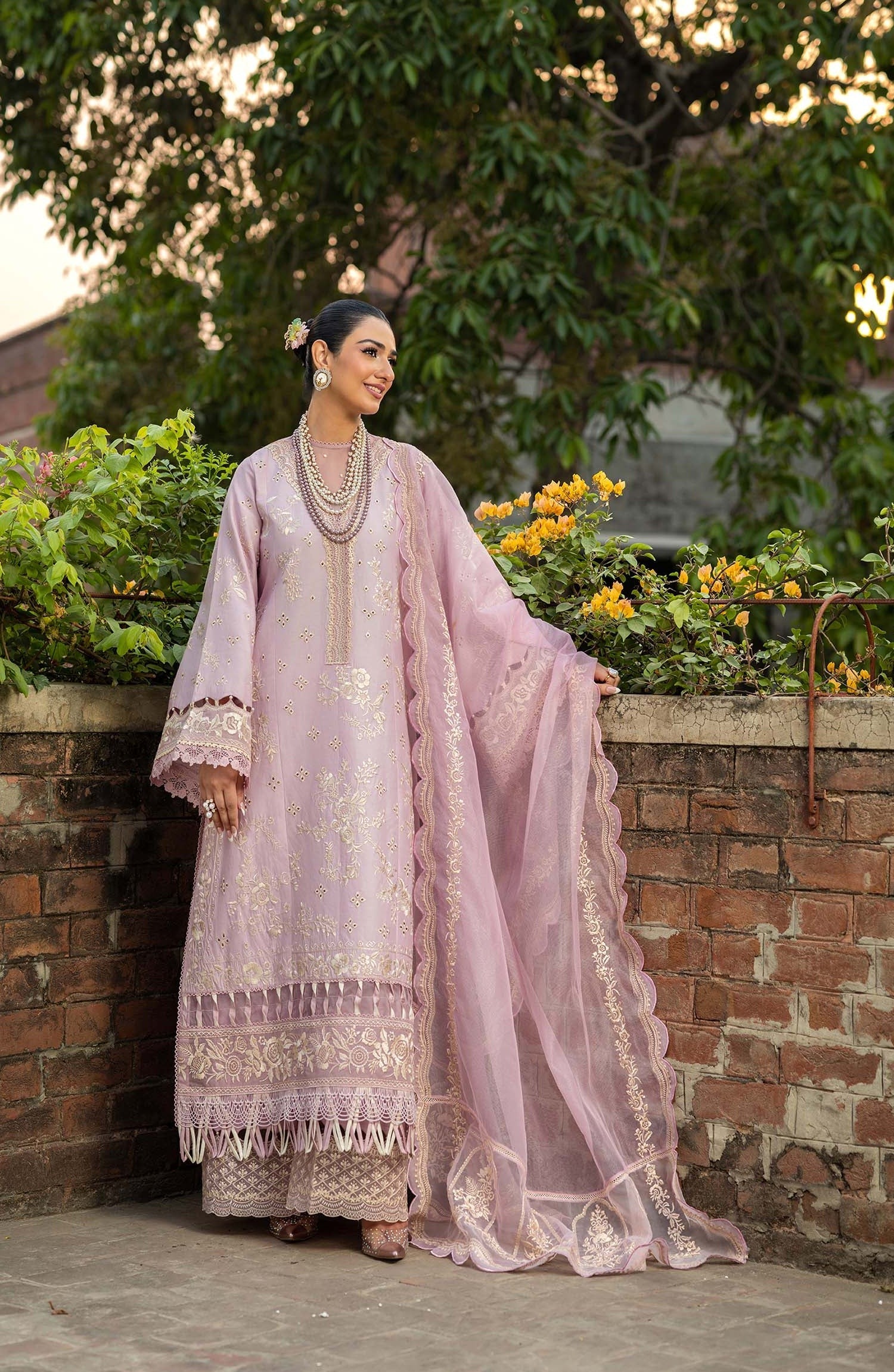 A woman wearing a light pink embroidered traditional outfit with a matching sheer dupatta, standing outdoors against a brick wall with greenery and flowers in the background.