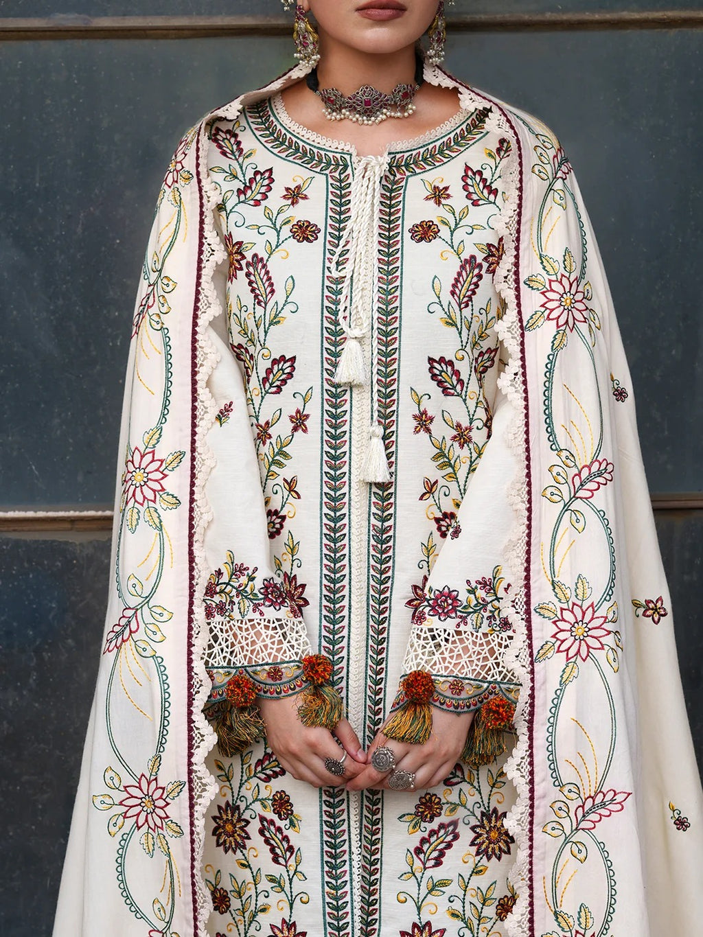 A close-up of a model wearing a heavily embroidered traditional outfit. The garment is off-white/cream with intricate floral and vine embroidery in green, red, orange, and yellow, featuring tassel ties and tigerskin cuff trim.