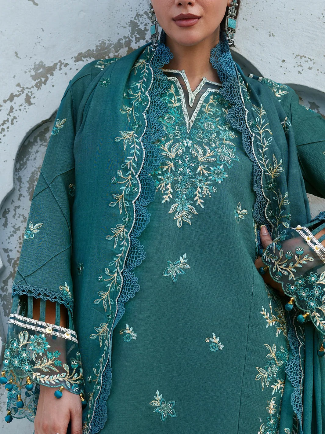 A close-up of a woman wearing a teal green traditional embroidered outfit with a matching scarf. The outfit features intricate floral embroidery in gold, white, and light blue, with lace trim and beaded cuffs.