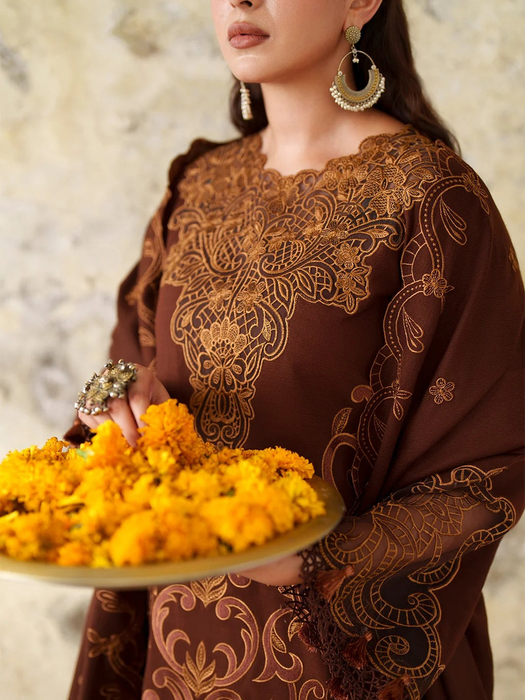 A woman wearing a brown embroidered kurta with intricate gold floral patterns, holding a plate of yellow marigold flowers against a textured stone wall background.