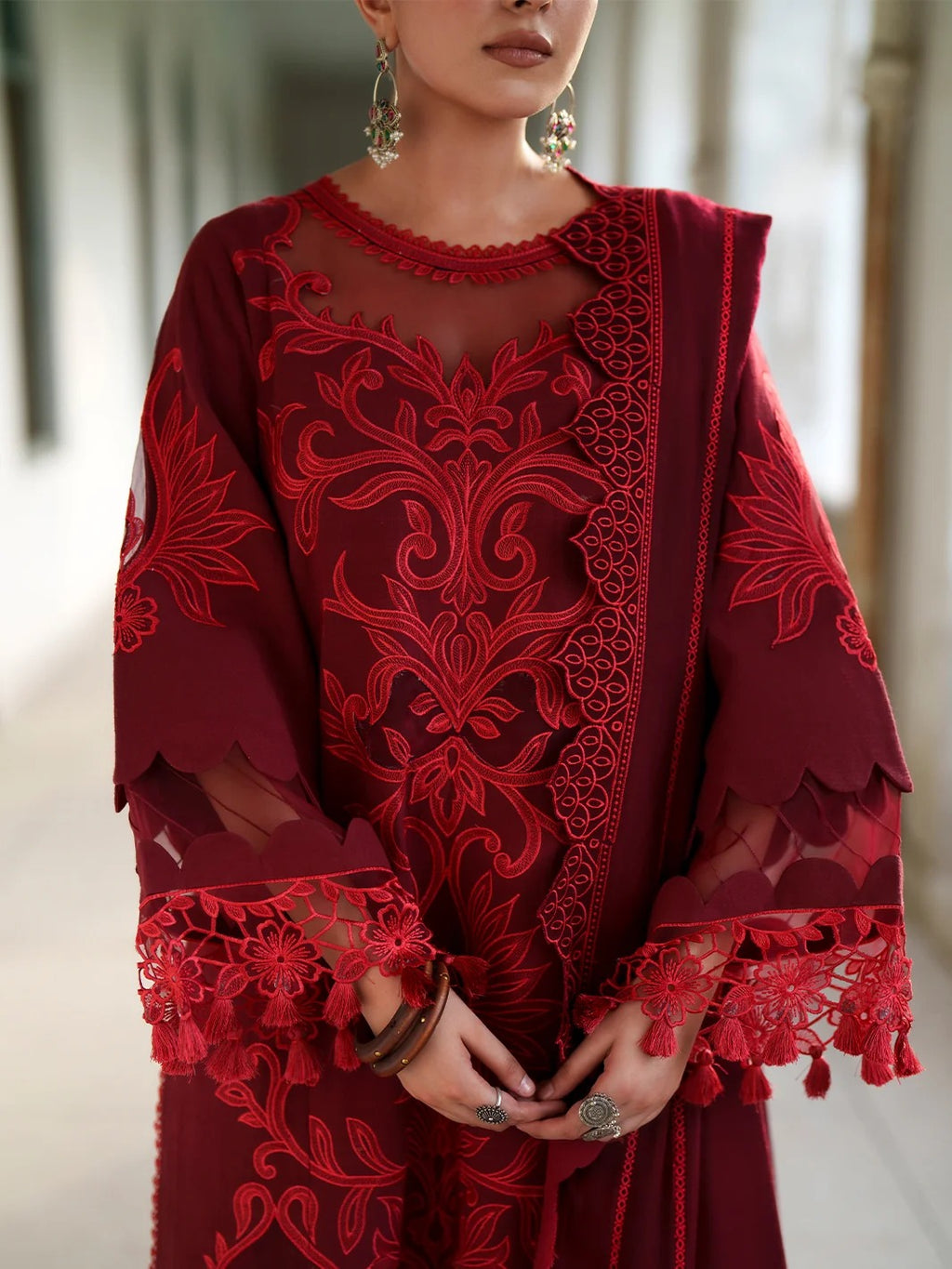 A woman wearing a deep red embroidered traditional outfit with sheer sleeves, lace trim, and matching dupatta, photographed in a softly blurred indoor setting.