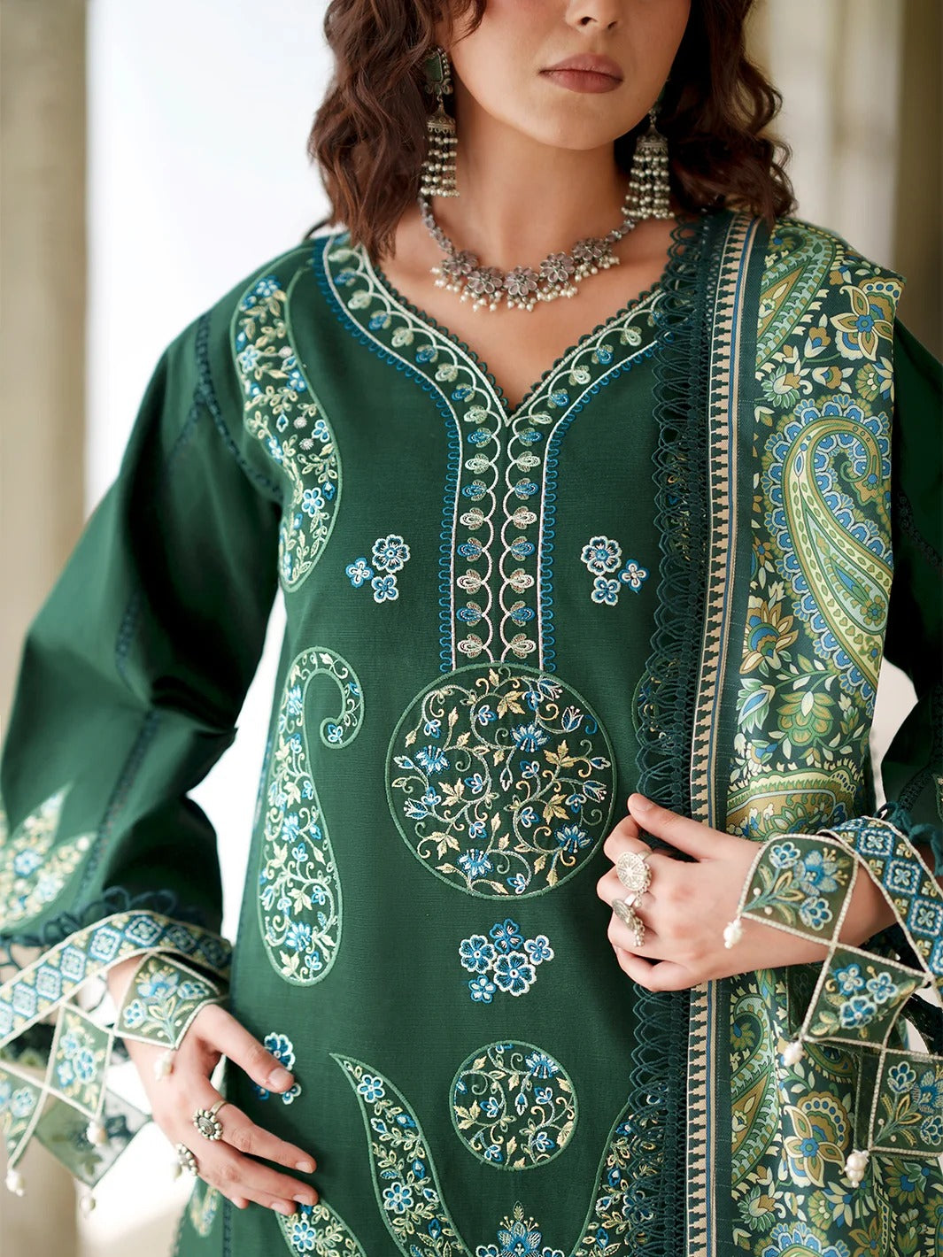A close-up of a woman wearing a dark green embroidered kurta with intricate paisley and floral patterns in light blue, beige, and white. She is accessorized with silver jewelry, including a statement necklace and earrings.