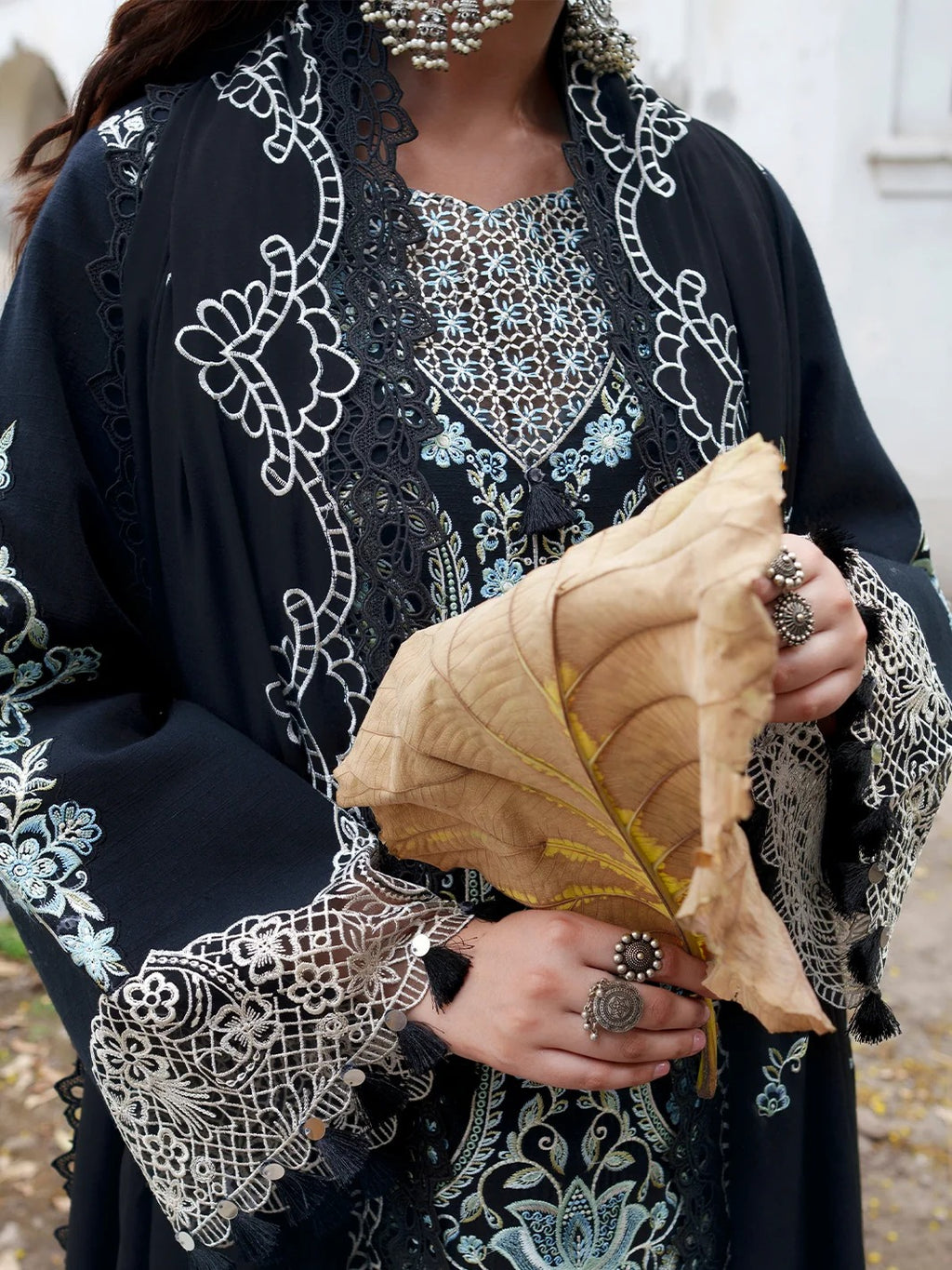 A close-up of a person wearing a black embroidered kaftan robe with white and light blue floral patterns, holding a dried tan leaf in both hands. The person is also wearing silver rings and a silver beaded necklace.