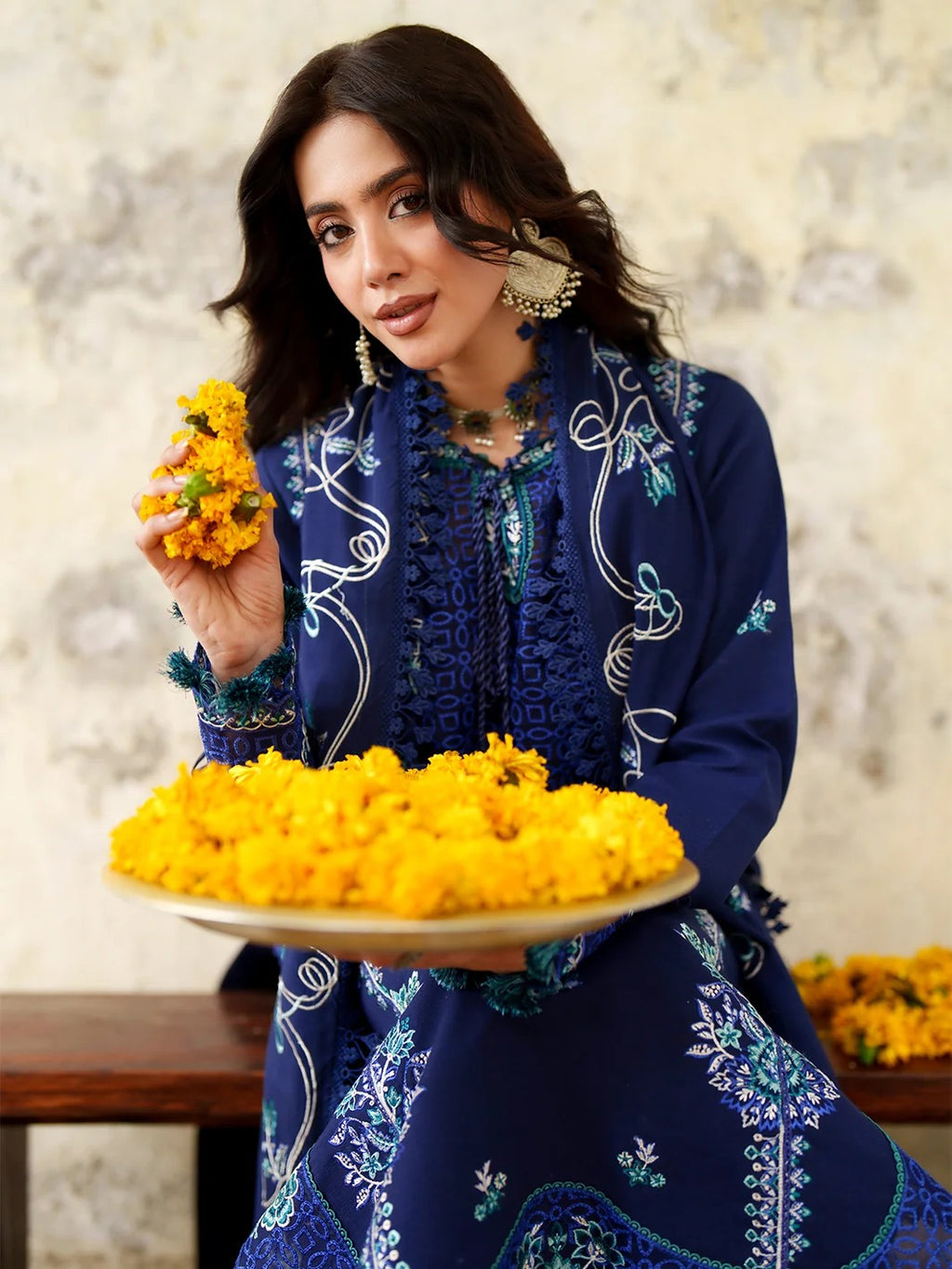 A woman wearing a navy blue embroidered kurta set holds a plate of yellow marigold flowers, with a matching yellow flower in her hand, set against a textured beige wall.