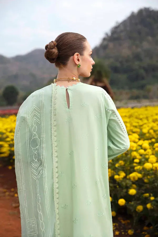 A woman wearing a mint green embroidered traditional outfit is photographed from behind in an outdoor field with yellow flowers and hills in the background.