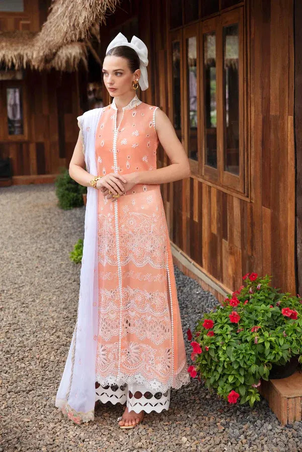 A woman stands outdoors in front of a wooden building wearing a peach embroidered traditional outfit with a white dupatta and white headscarf. The setting includes gravel ground, potted red flowers, and a thatched roof in the background.