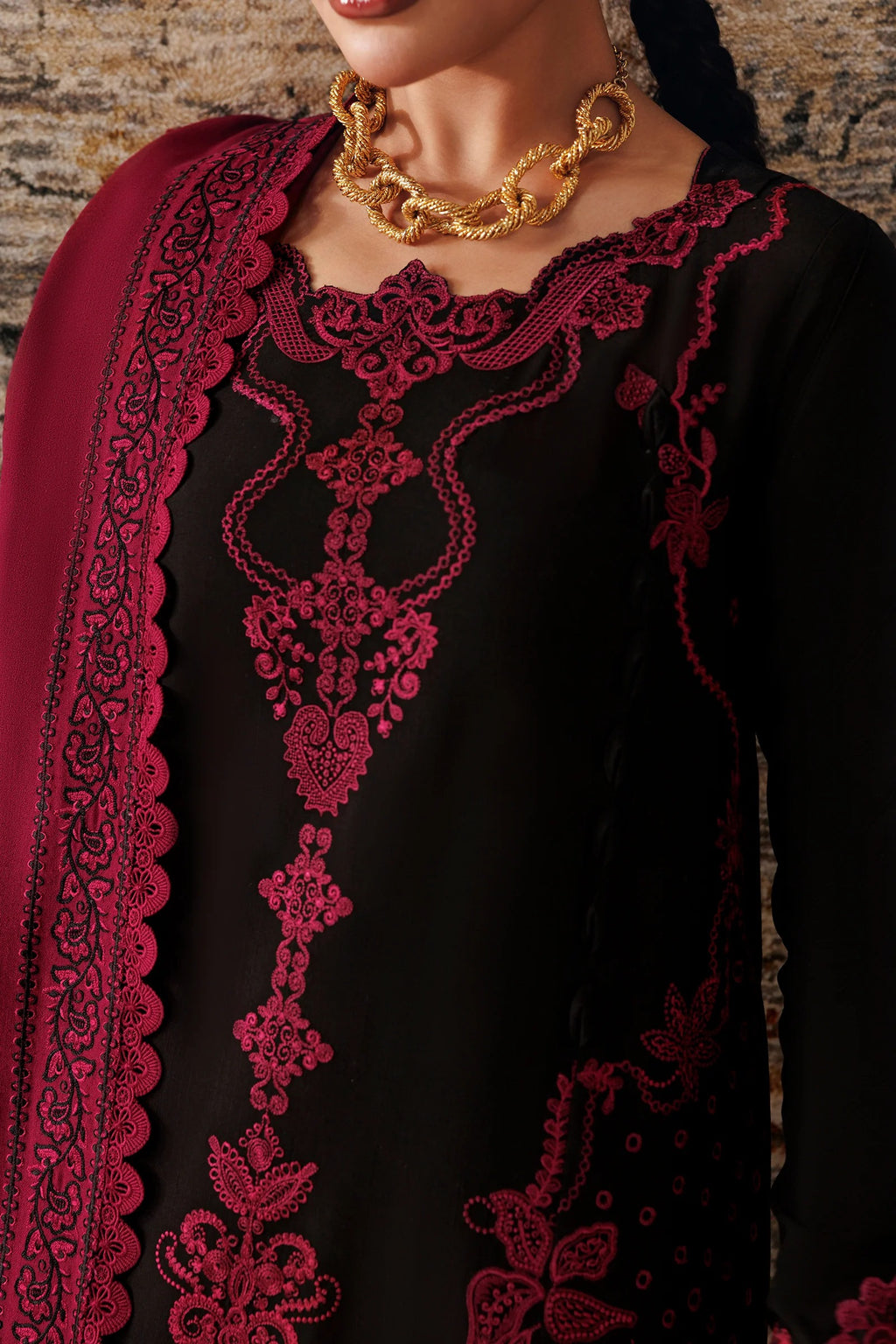A close-up of a black traditional outfit with deep red floral embroidery and a matching red embroidered border, worn by a model styled with a chunky gold necklace.