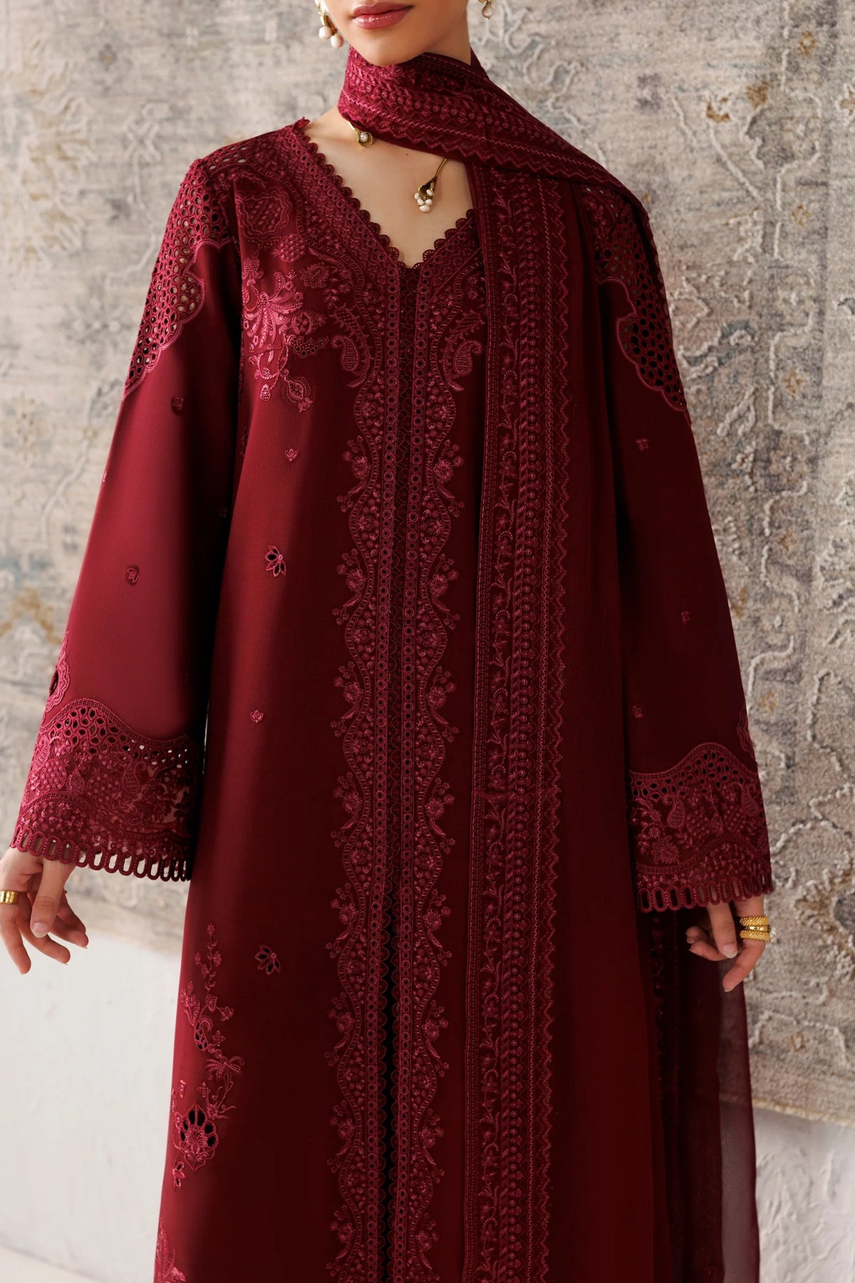 A woman wearing a long, deep burgundy embroidered dress with a matching neck scarf, shown from the neck down against a textured gray wall.