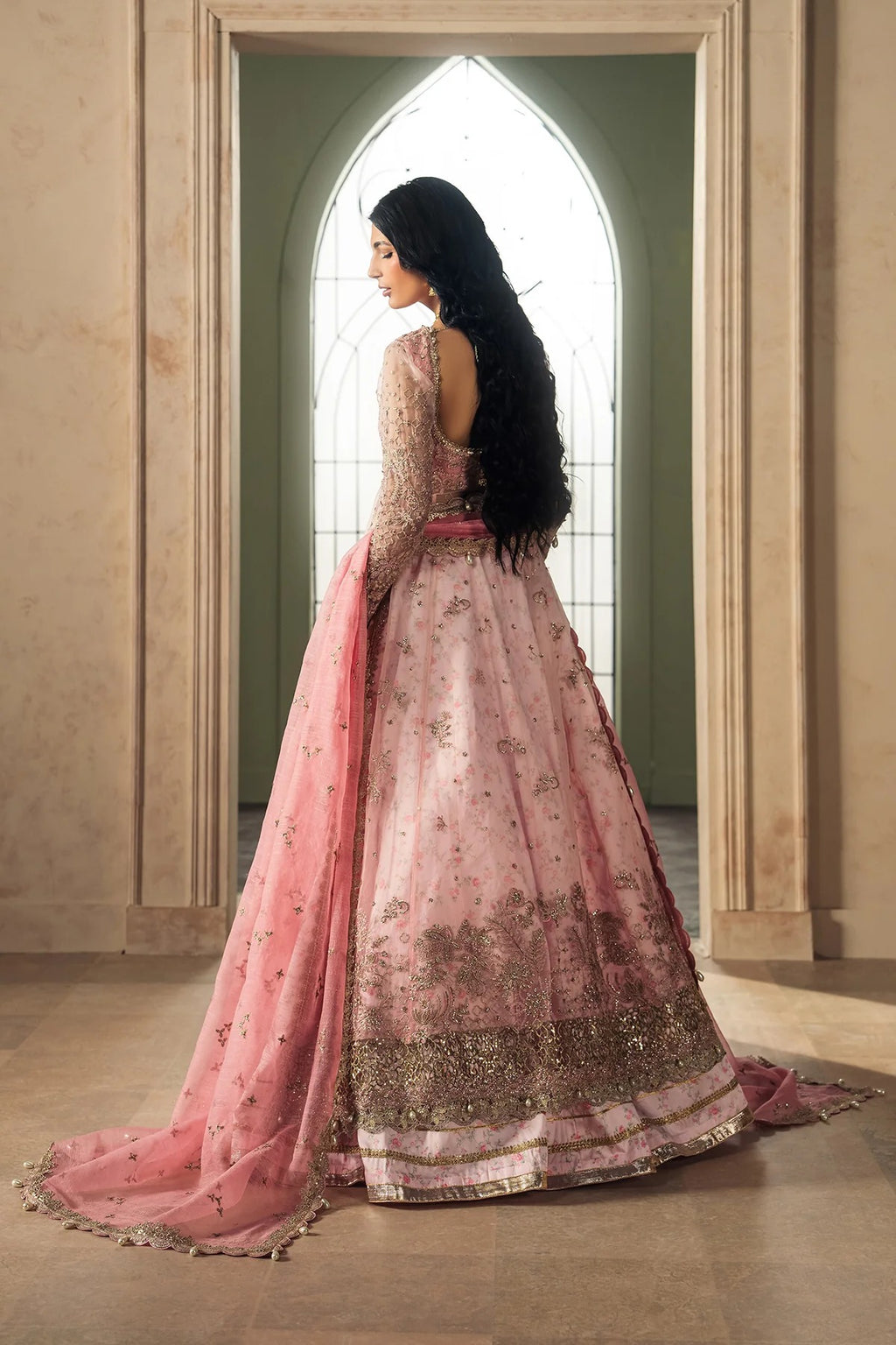 A woman wearing a pink embroidered traditional gown with a long train and sheer dupatta, standing in profile in front of an arched window in a beige interior.