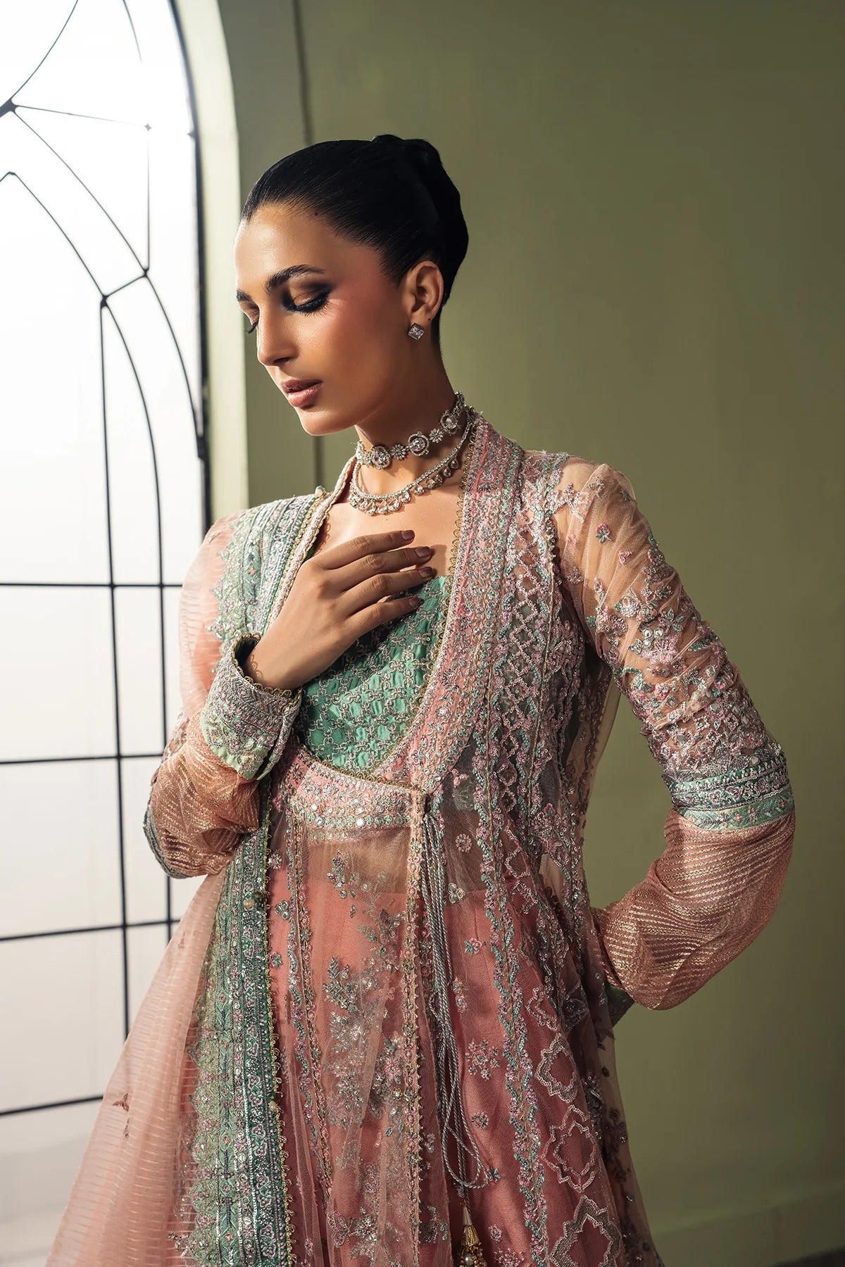 A woman models an ornate traditional outfit with a dusty pink embroidered outer layer, a mint green inner dress, and a matching sheer pink dupatta, set against a soft olive-green wall and a window frame.