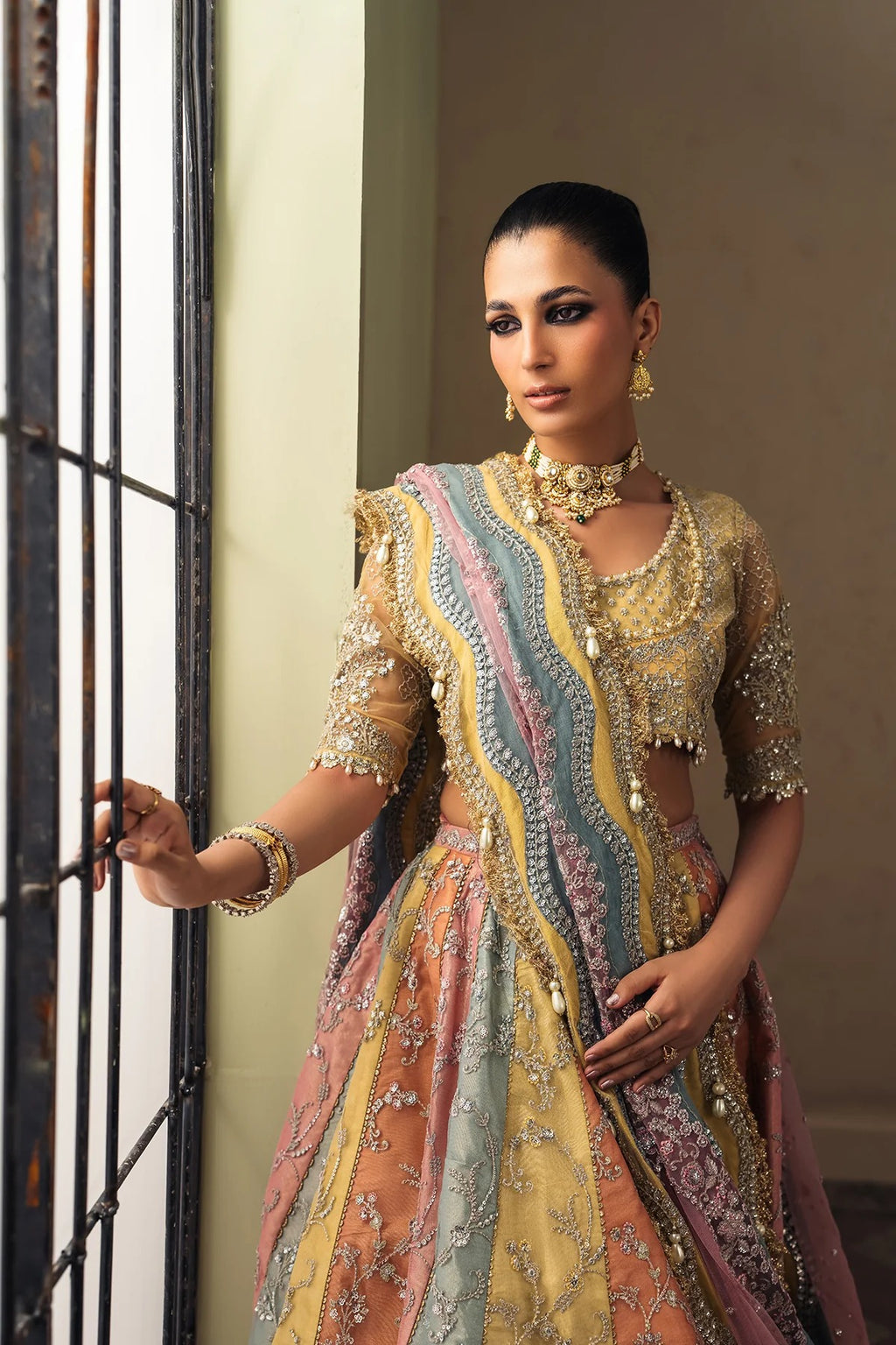 A woman models a traditional Indian lehenga choli set with intricate gold embroidery and multicolor panels of pink, blue, and yellow, standing next to a window.