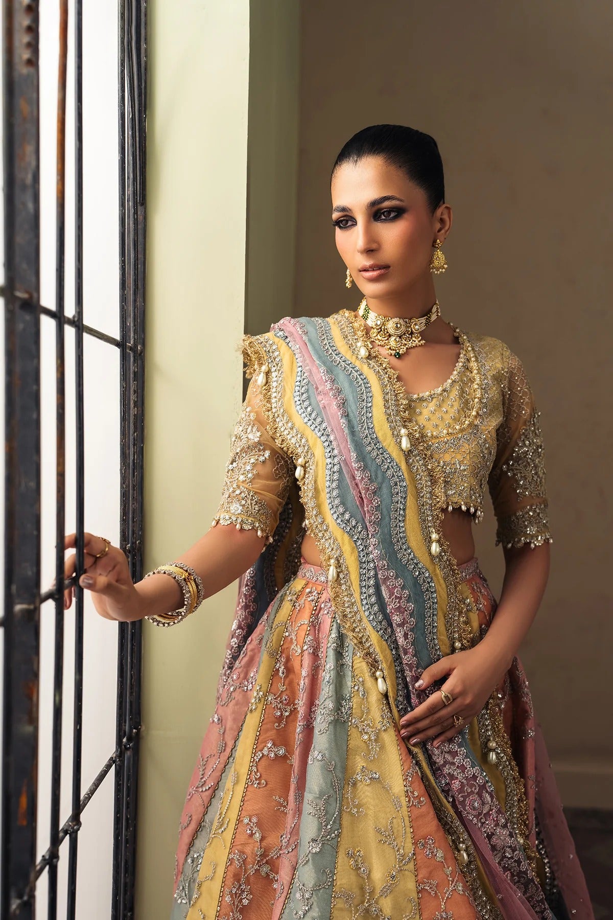 A woman models a traditional Indian lehenga choli set with intricate gold embroidery and multicolor panels of pink, blue, and yellow, standing next to a window.