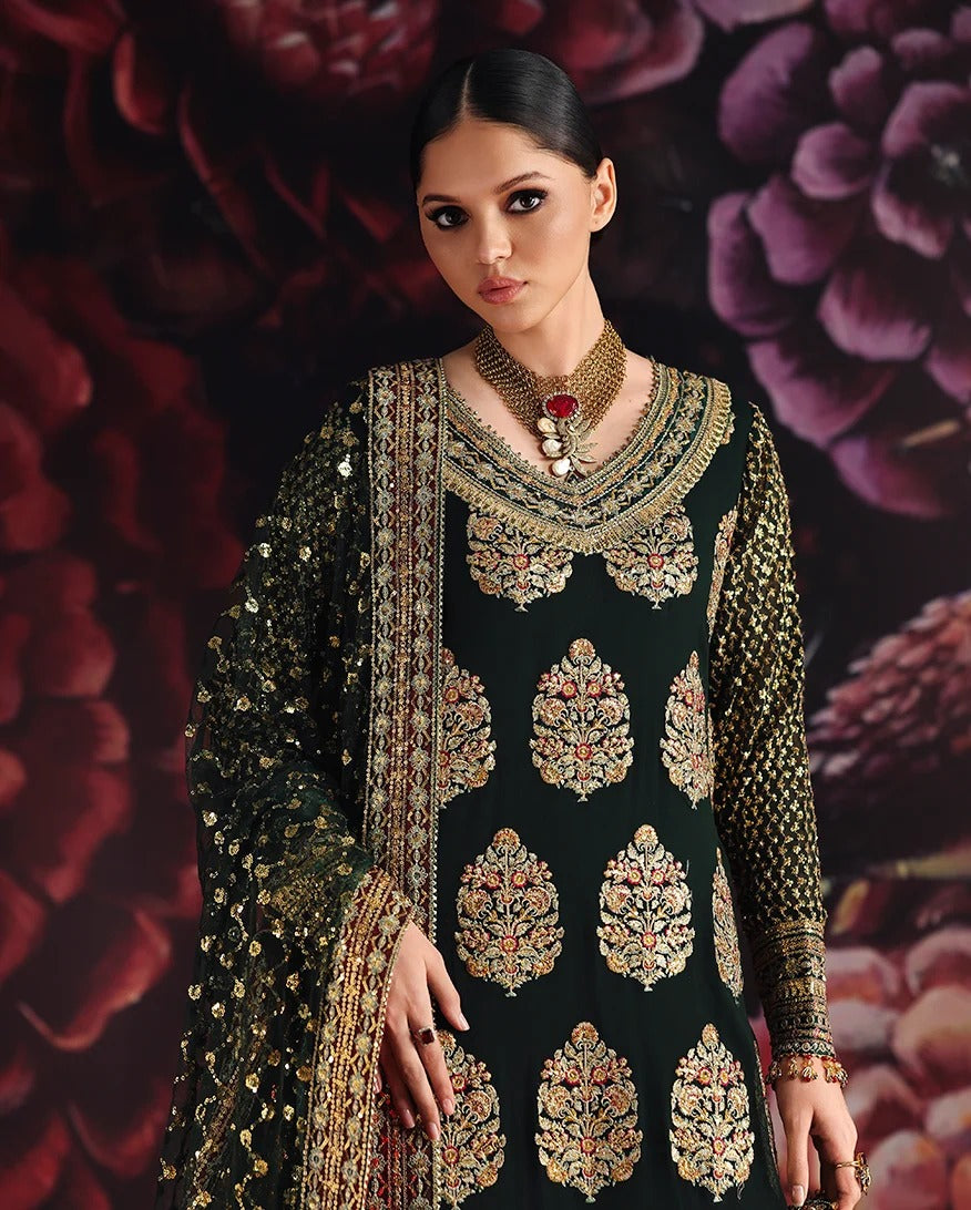A woman modeling a black traditional embroidered outfit with ornate gold, red, and green motifs, paired with a sheer embroidered dupatta, set against a dark floral background.