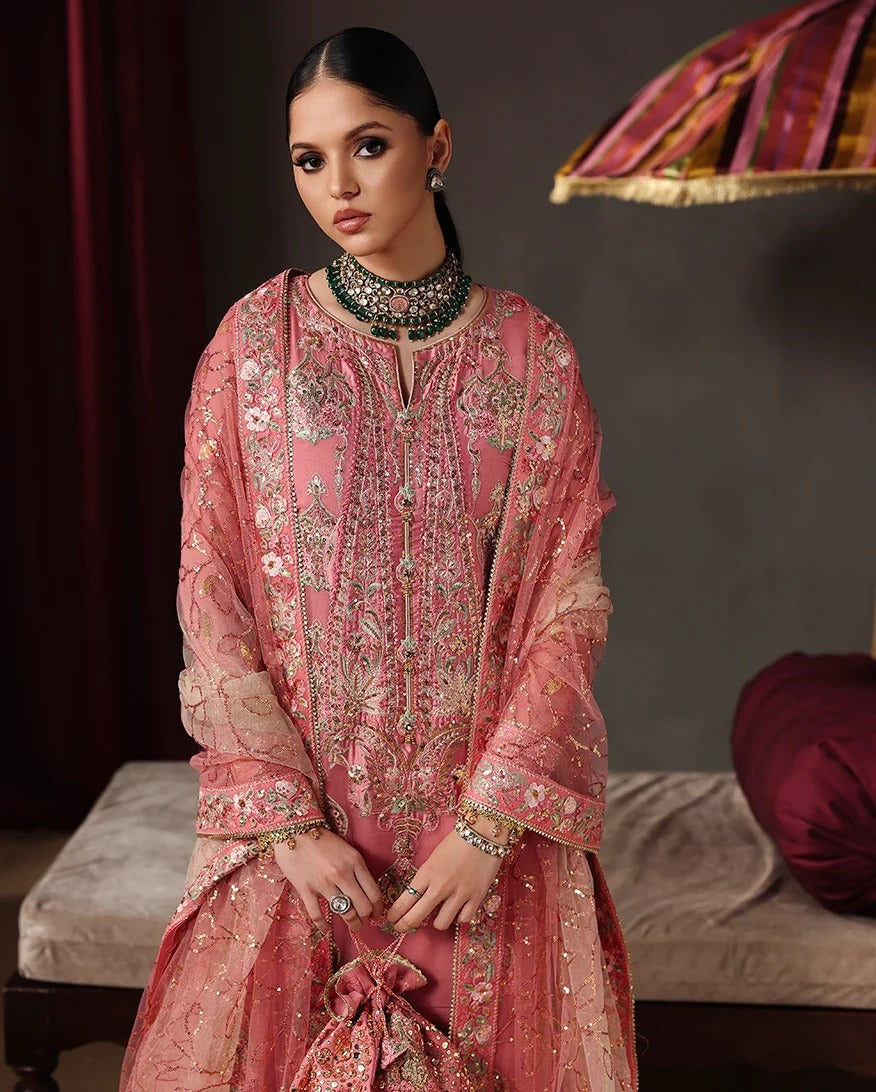 A woman models a traditional pink embroidered kurta set with a matching dupatta in a dimly lit, elegant room. The outfit features intricate gold, silver, and green mirror work embroidery on a sheer pink mesh fabric.