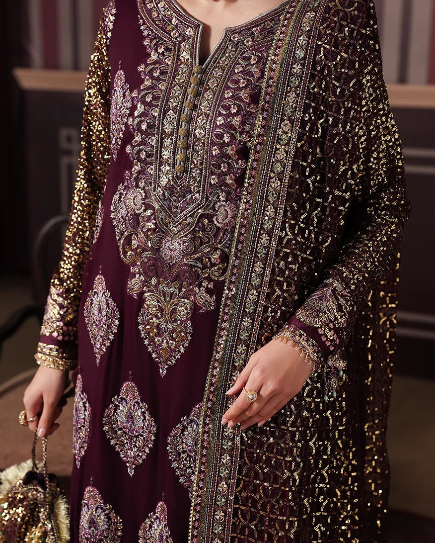 A close-up of a woman wearing a deep maroon traditional embroidered outfit with intricate gold and silver mirror work, sequins, and embroidery. The garment features a deep V-neckline and long sleeves with dense decorative patterns.