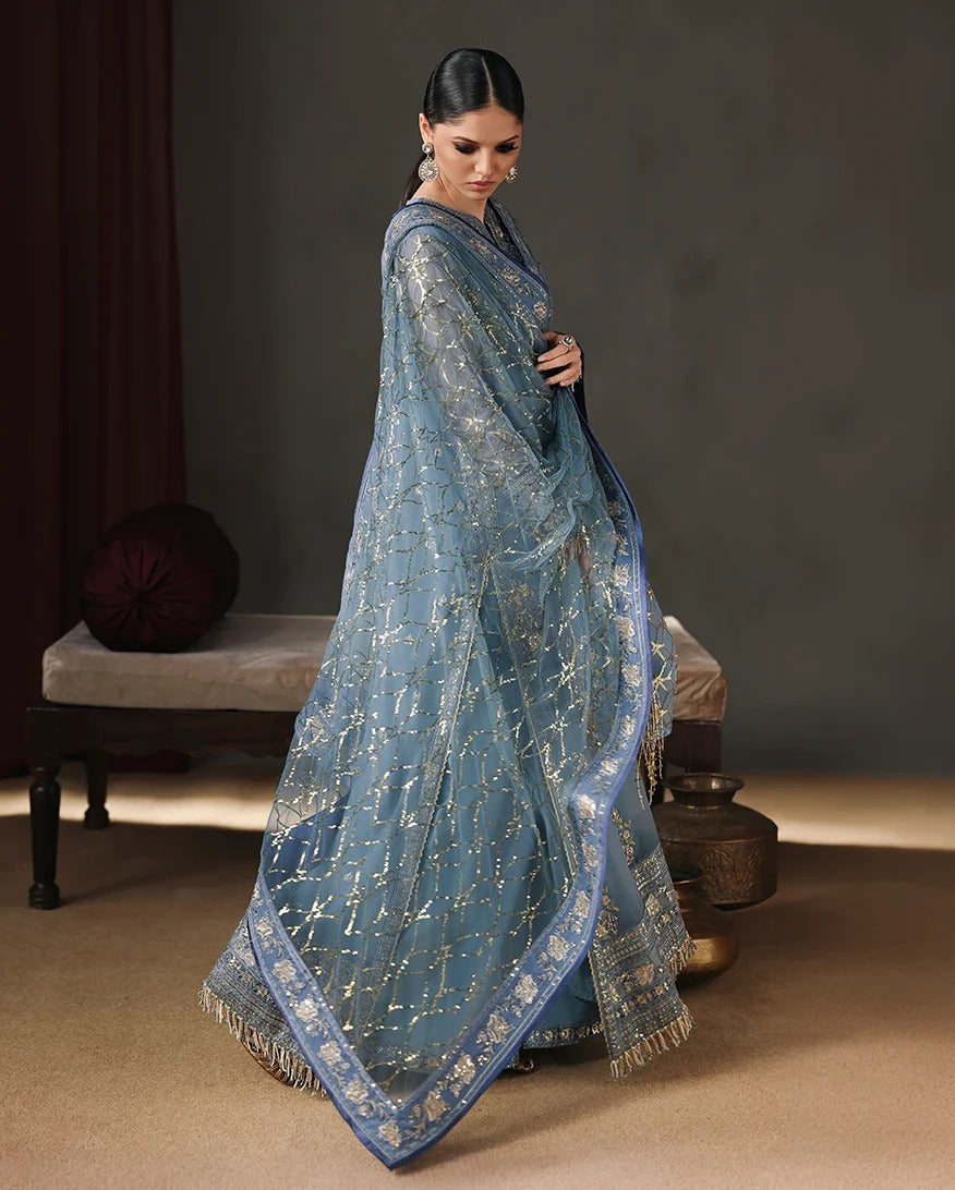 A woman models a light blue saree with intricate gold zari embroidery and a matching blue border, posed in a styled studio setting with a bench and decorative pot in the background.