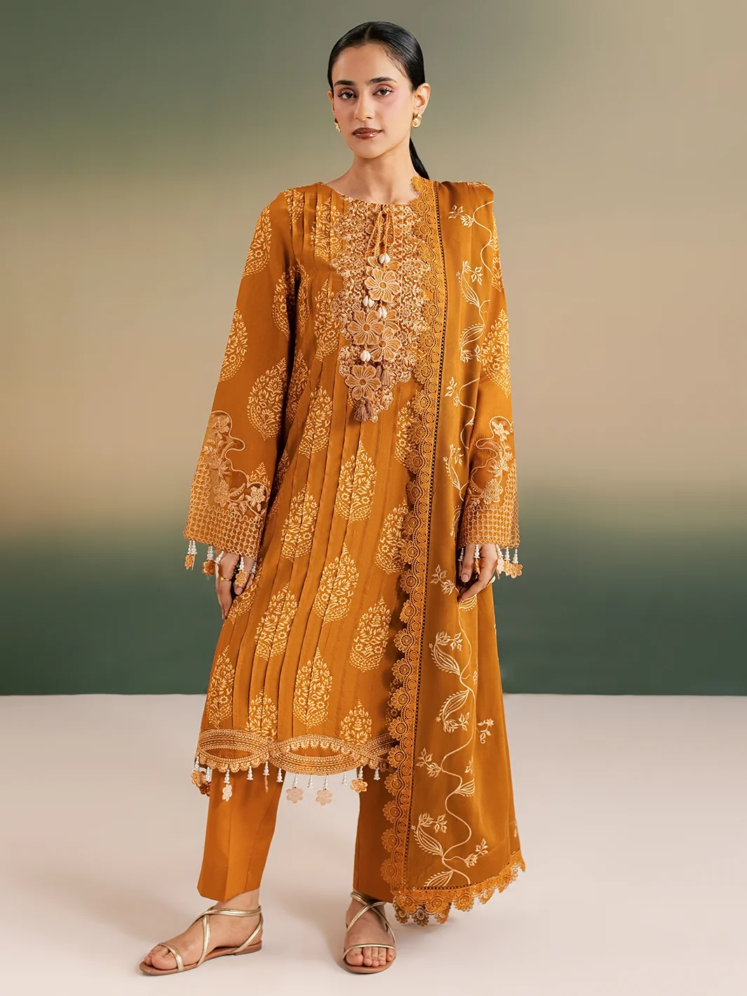 A woman modeling a traditional South Asian outfit consisting of a mustard yellow embroidered kurta, matching trousers, and a coordinating dupatta, set against a neutral studio backdrop.