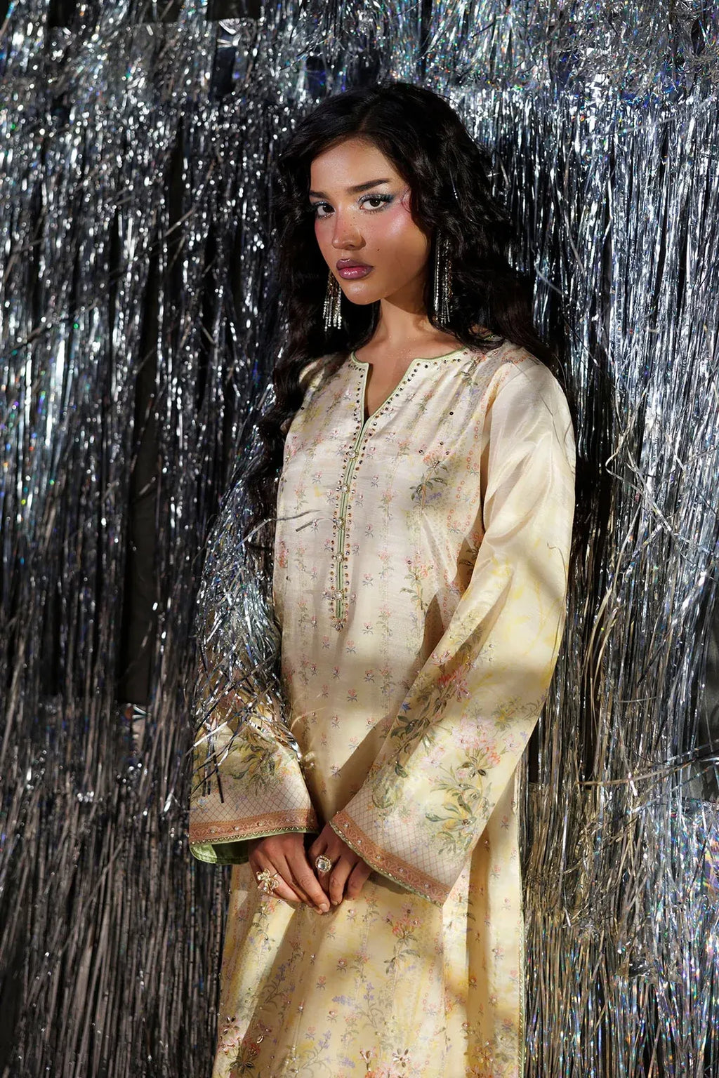 A woman wearing a cream-colored embroidered tunic dress with floral patterns and silver embellishments, standing in front of a shimmering silver tinsel backdrop.