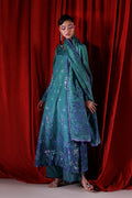 A model wearing a teal blue embroidered traditional outfit with a matching dupatta, standing in front of a red velvet curtain backdrop.
