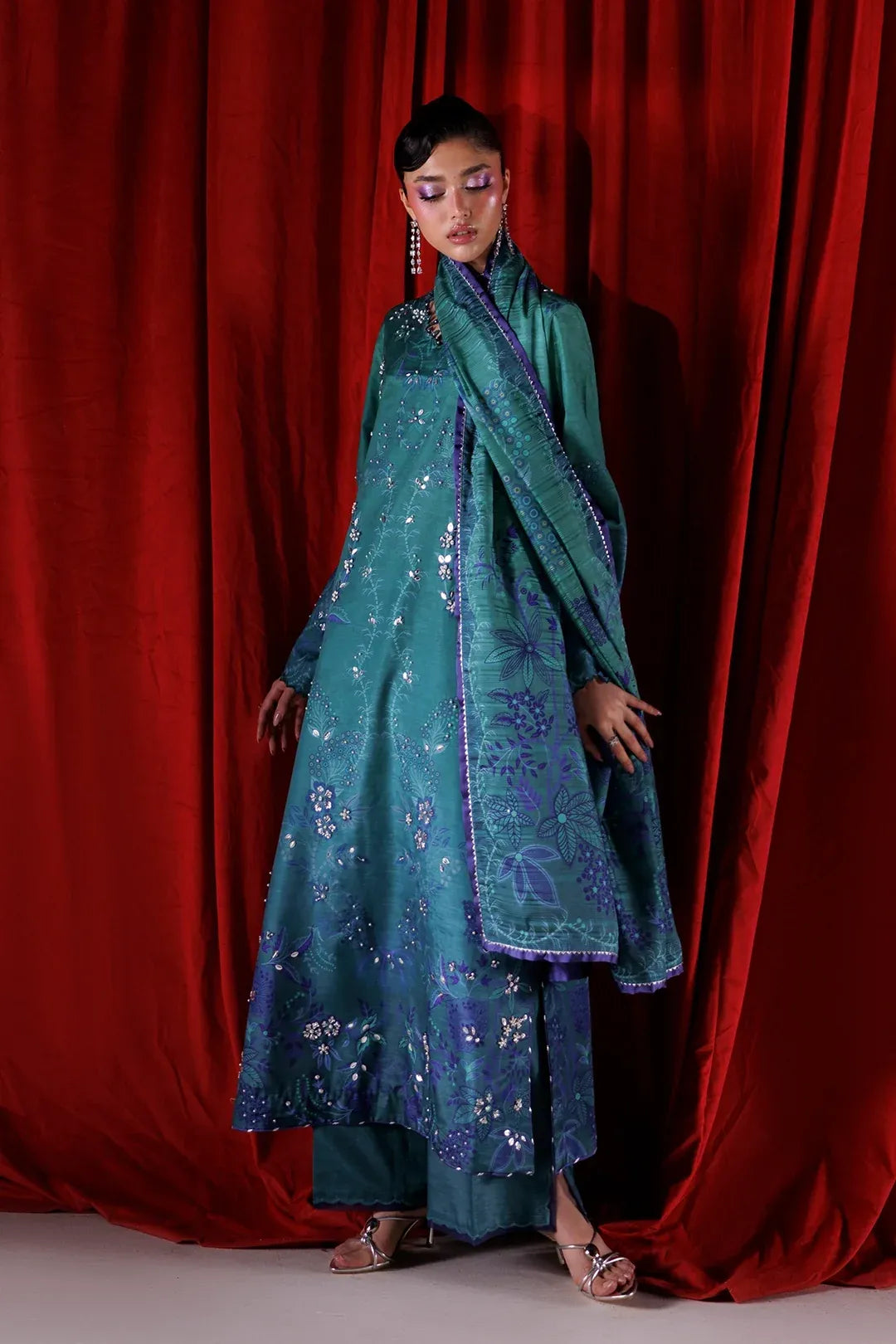 A model wearing a teal blue embroidered traditional outfit with a matching dupatta, standing in front of a red velvet curtain backdrop.