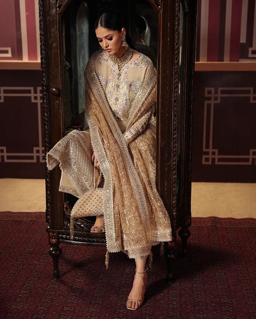 A woman seated in an ornate dark wooden cabinet wearing a heavily embellished beige and gold traditional outfit with a matching dupatta, photographed in a richly decorated interior with patterned walls and a red carpet.