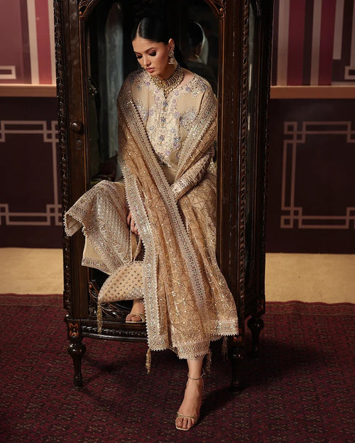 A woman seated in an ornate dark wooden cabinet wearing a heavily embellished beige and gold traditional outfit with a matching dupatta, photographed in a richly decorated interior with patterned walls and a red carpet.