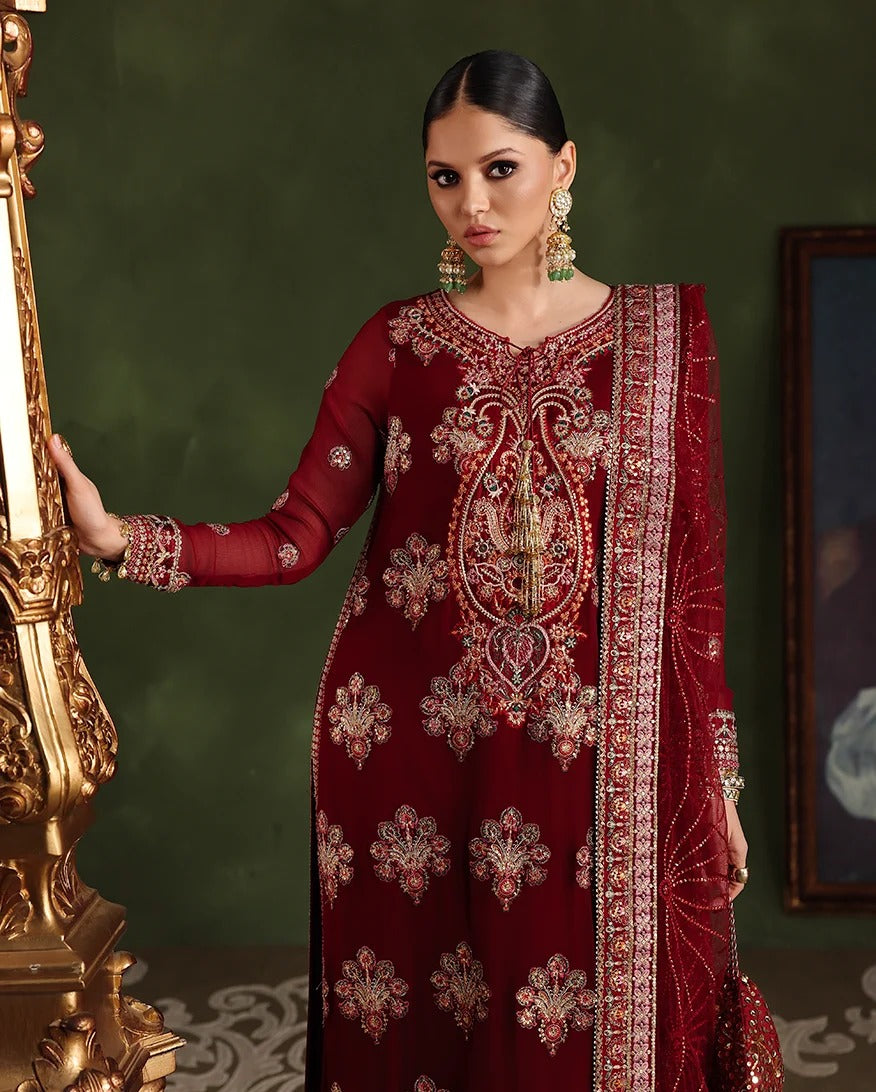 A woman models a deep maroon red traditional embroidered kurta set with a matching dupatta, featuring intricate gold and silver mirror work embroidery. She is posed next to an ornate gold pillar against a dark green wall.