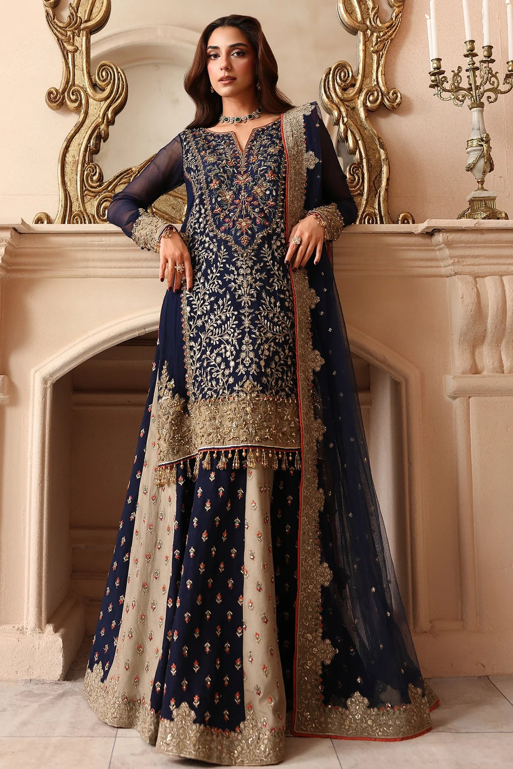 A woman models a traditional Indian salwar kameez suit with a navy blue and beige kurta, heavily embroidered with gold and silver threadwork, paired with a matching navy blue dupatta, set against an ornate beige fireplace background.