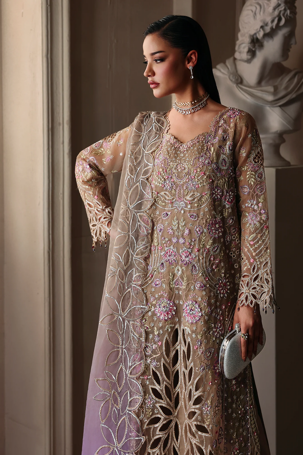 A woman models a traditional embroidered outfit with a beige and lavender base, heavily decorated with silver, pink, and green floral embroidery. She is posed in front of a classical stone bust in a warm, elegant interior.