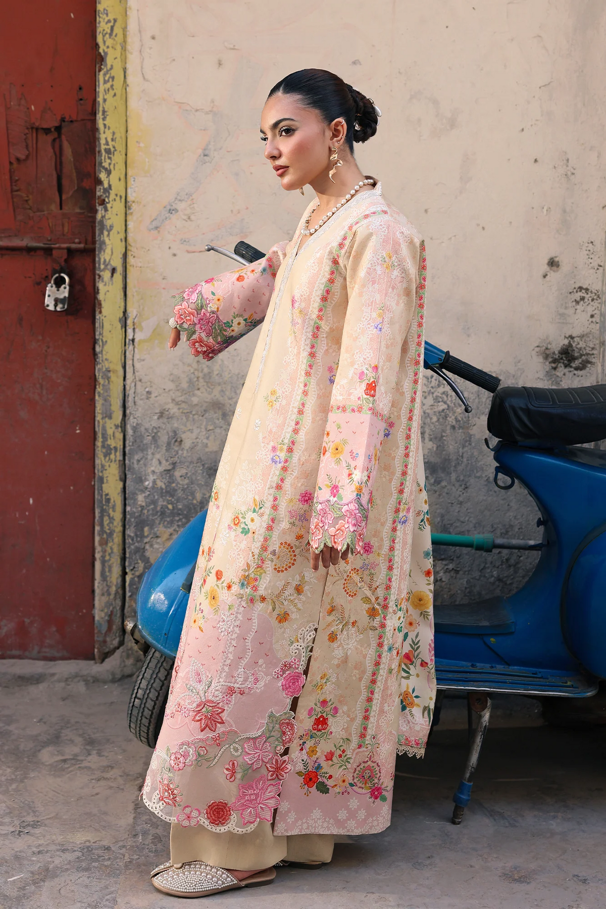 A woman models a traditional embroidered kurta in a light beige and pink colorway, standing next to a blue scooter against a weathered wall.
