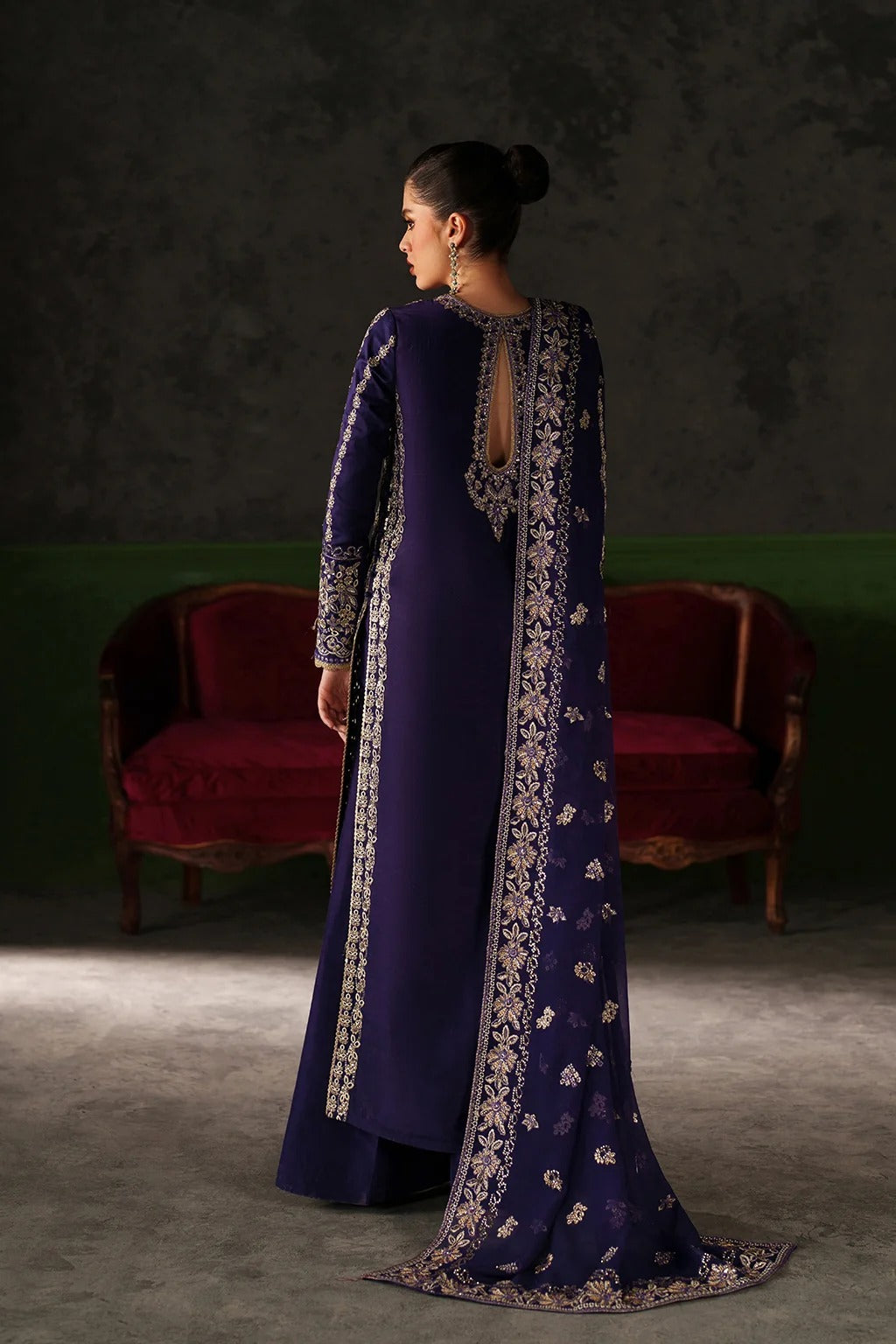 A woman stands with her back to the camera wearing a deep purple traditional outfit with ornate silver embroidery and a matching embroidered dupatta, posed in front of a dark textured wall and red velvet chairs.