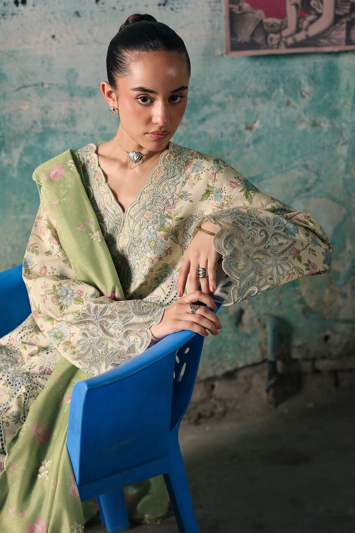 A woman wearing a traditional embroidered outfit with a light green dupatta sits on a blue plastic chair against a textured, peeling teal wall.