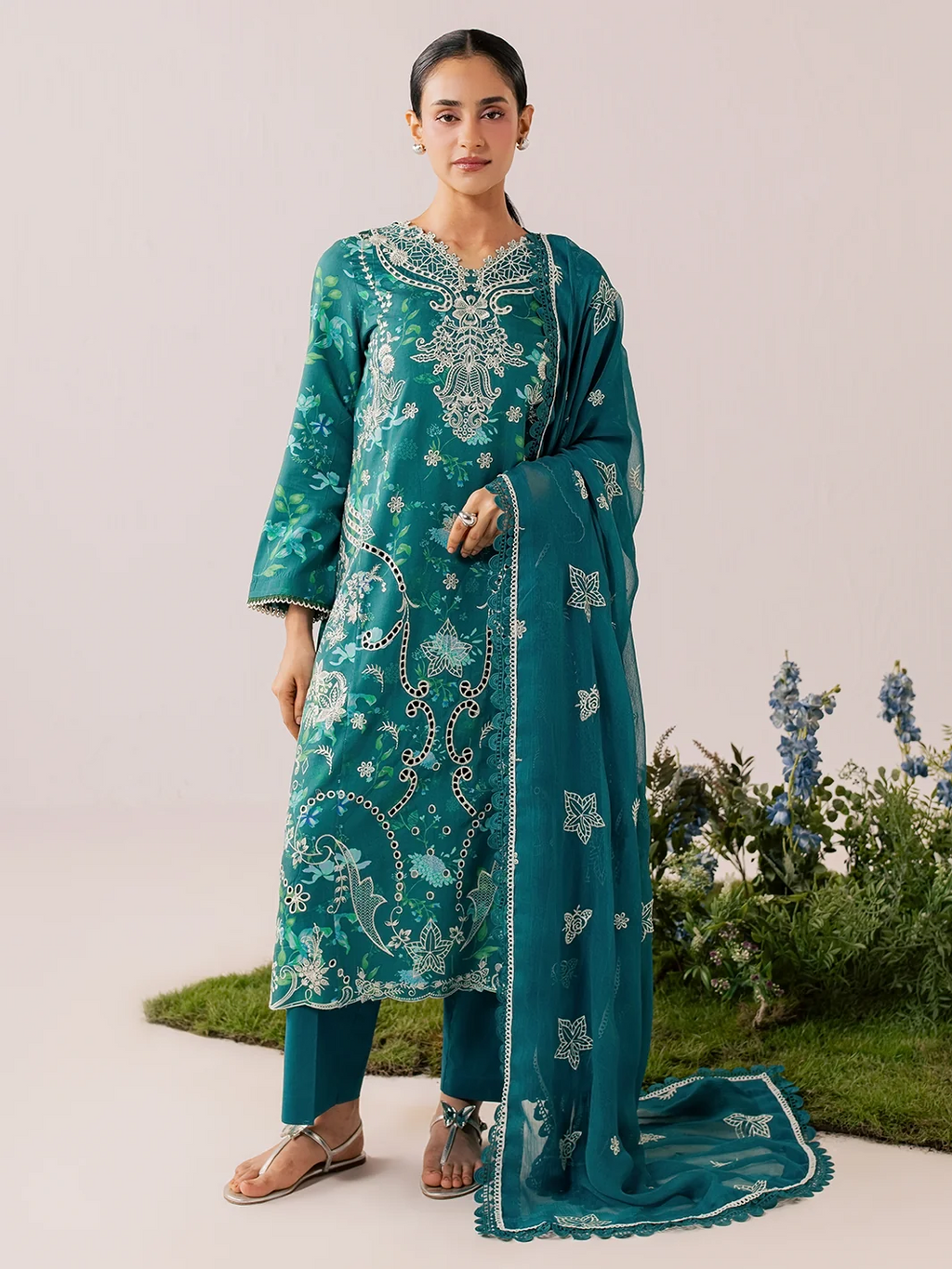 A woman modeling a teal green traditional Indian salwar kameez suit with intricate silver embroidery, matching trousers, and a sheer dupatta, set against a neutral studio background with decorative plants.