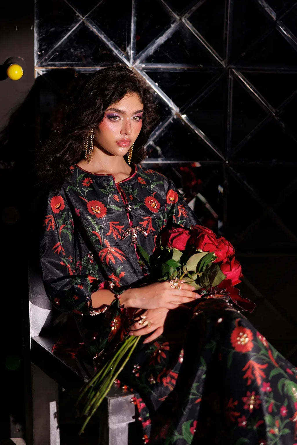 A model wearing a black embroidered dress with red floral patterns sits holding a bouquet of red roses in front of a dark geometric background.