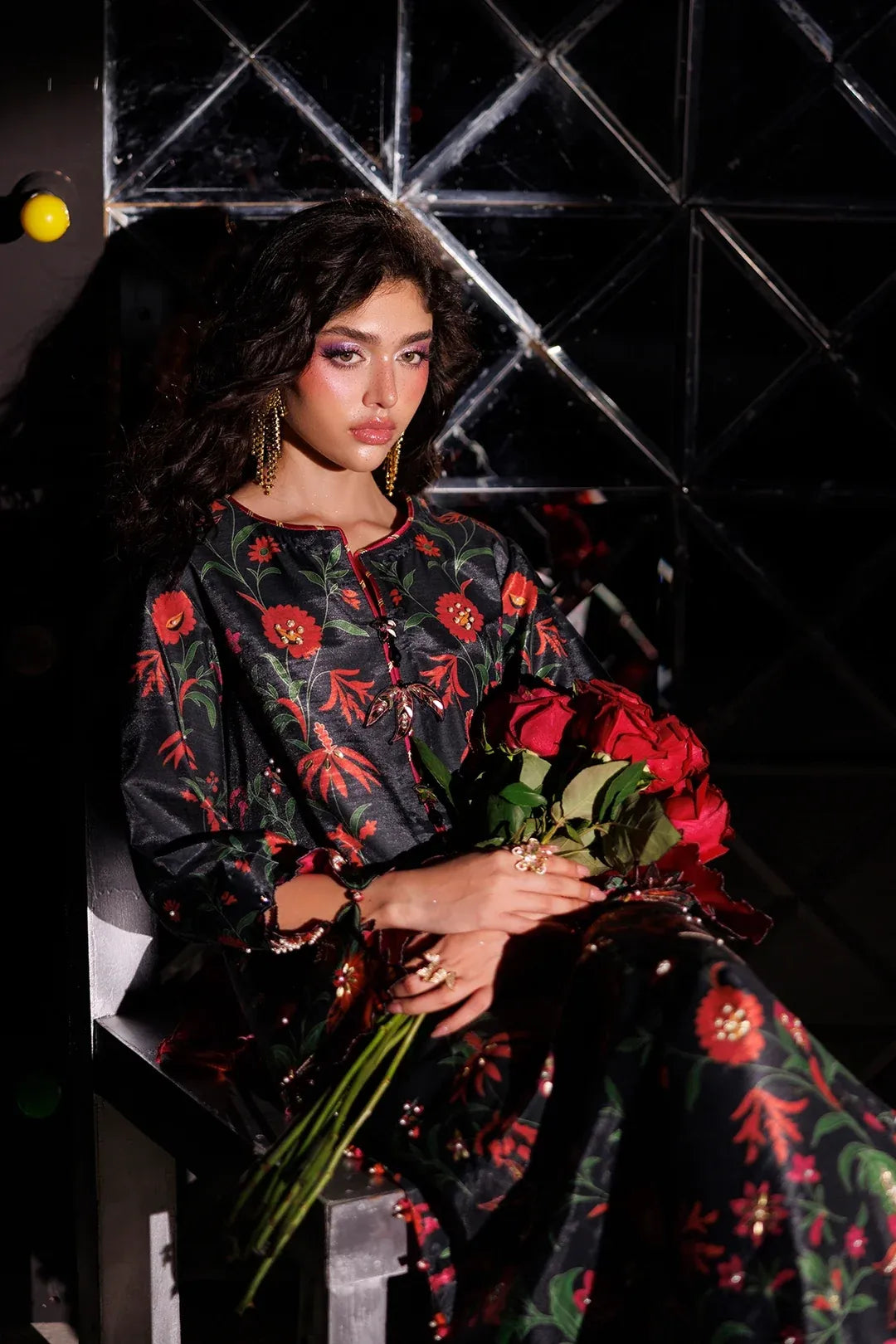 A model wearing a black embroidered dress with red floral patterns sits holding a bouquet of red roses in front of a dark geometric background.