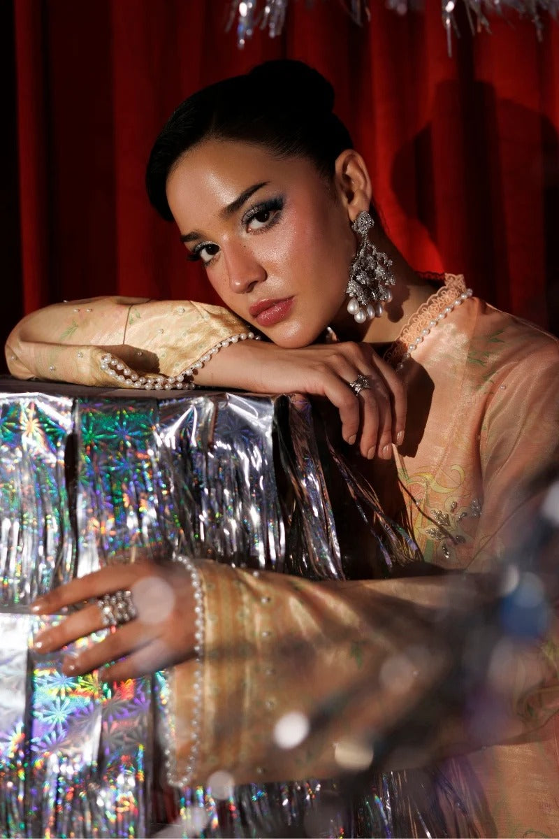 A woman in a sheer peach embroidered traditional-style top leans over a reflective holographic surface, wearing pearl necklaces, rings, and large silver earrings against a red curtain background.